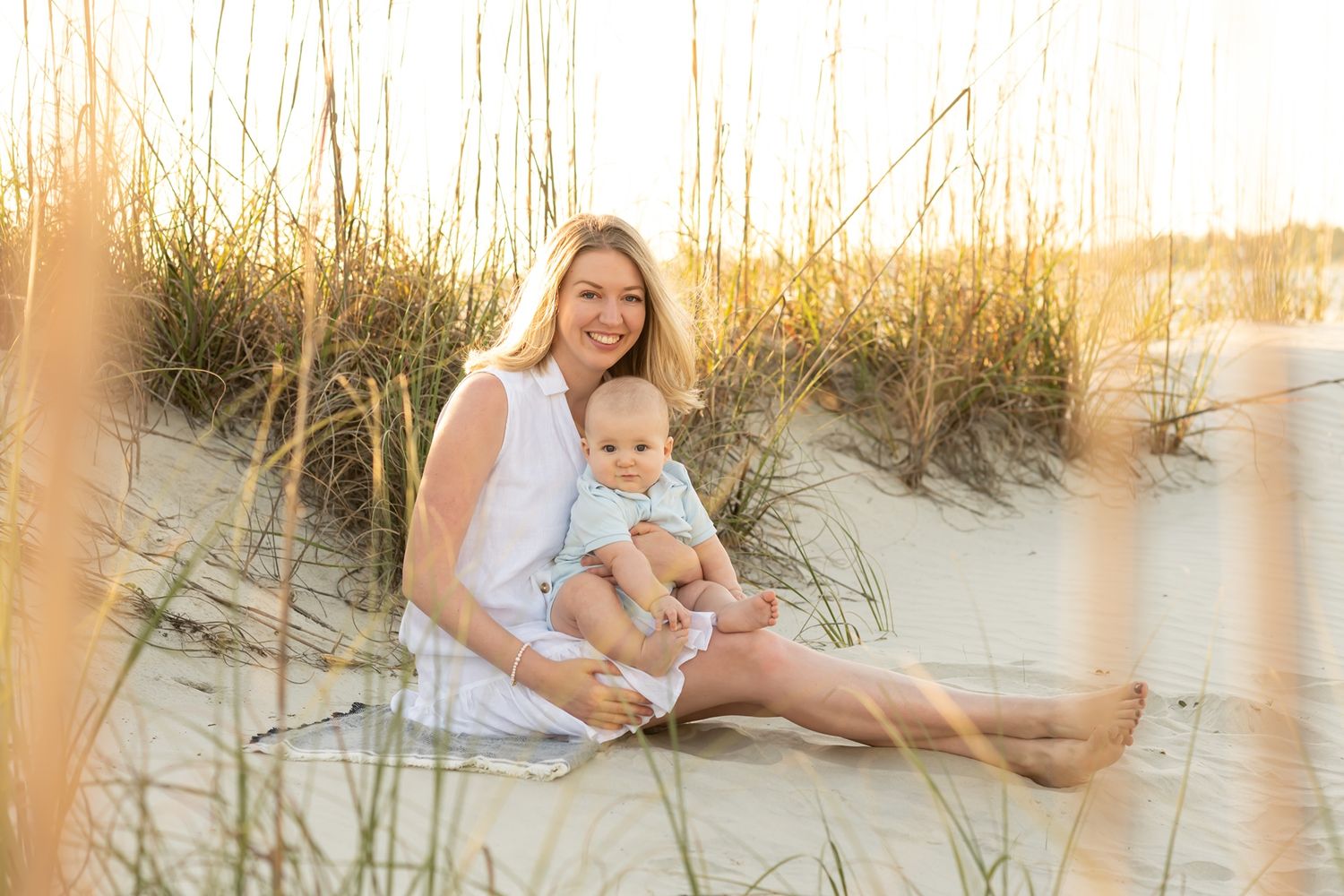 Two figures sit together in beach grass during a golden sunset, creating a serene coastal portrait scene.