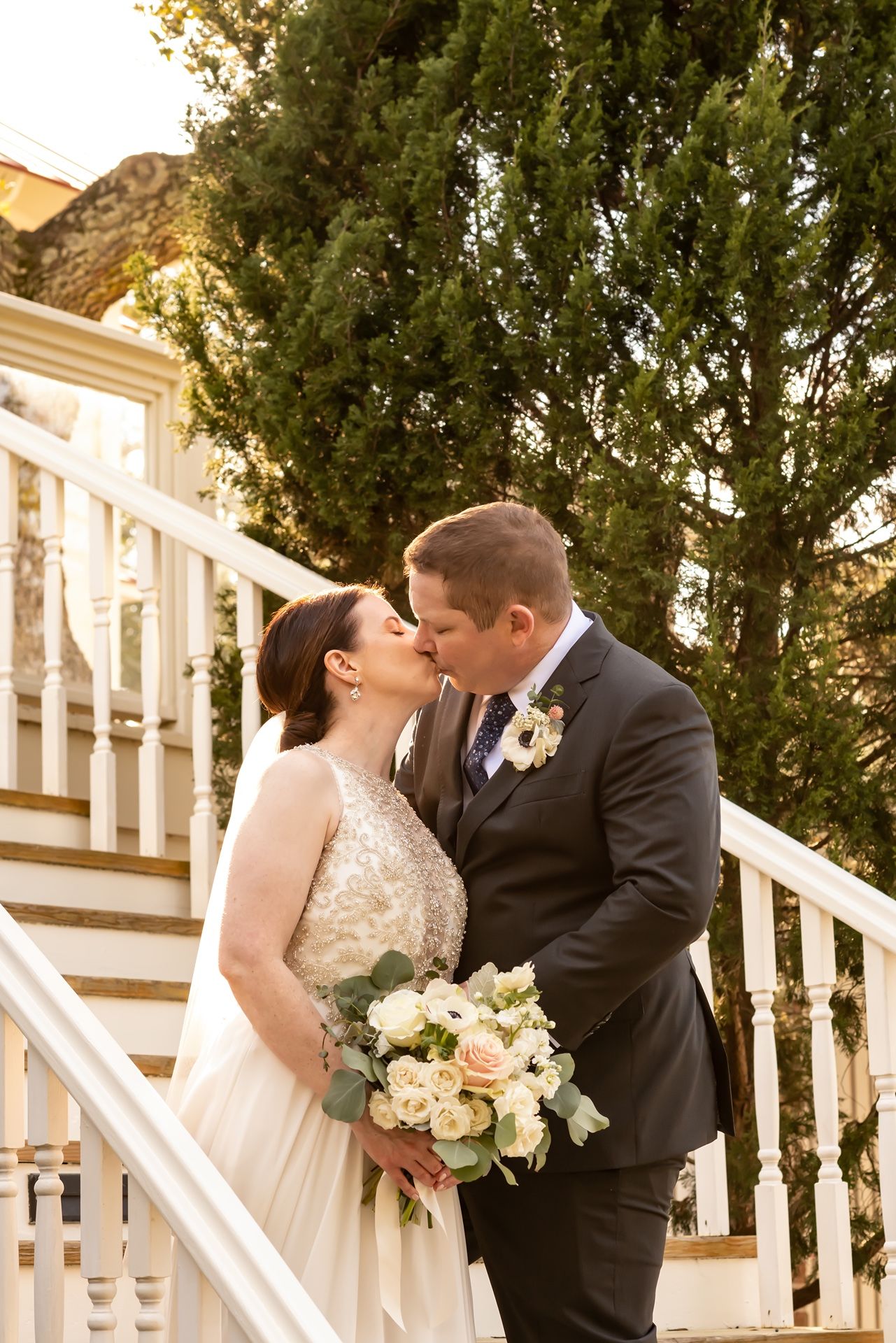 Couple shares romantic kiss on white staircase with lush greenery behind them, bride holding elegant white bouquet.