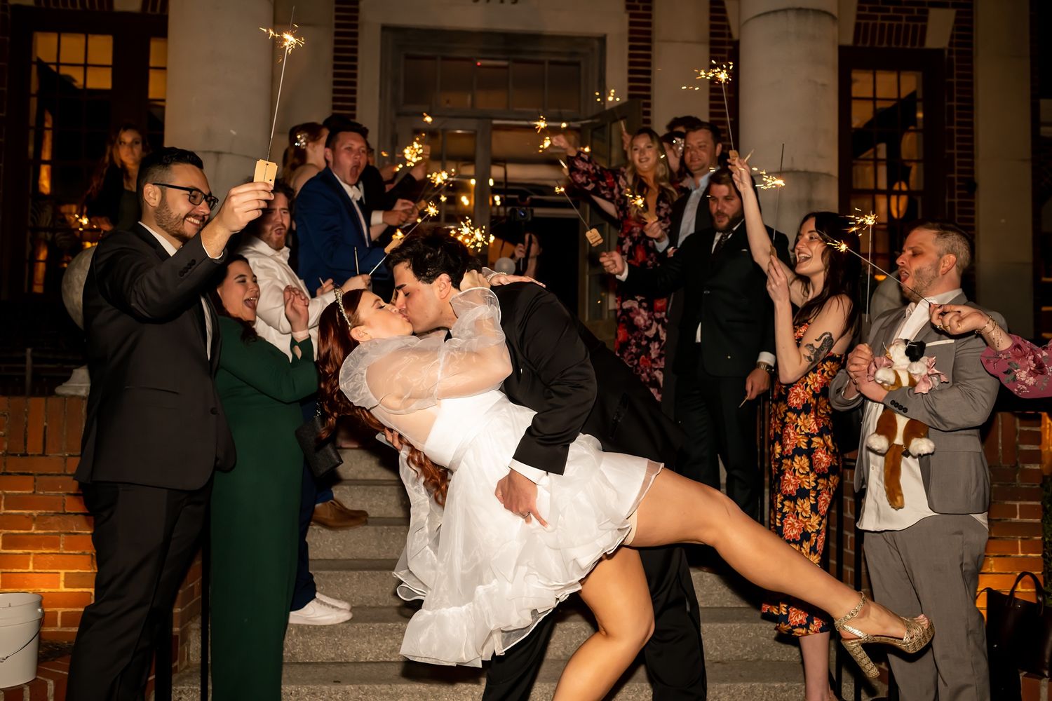 A couple shares a celebratory dip kiss while surrounded by guests holding sparklers outside an elegant venue at night.