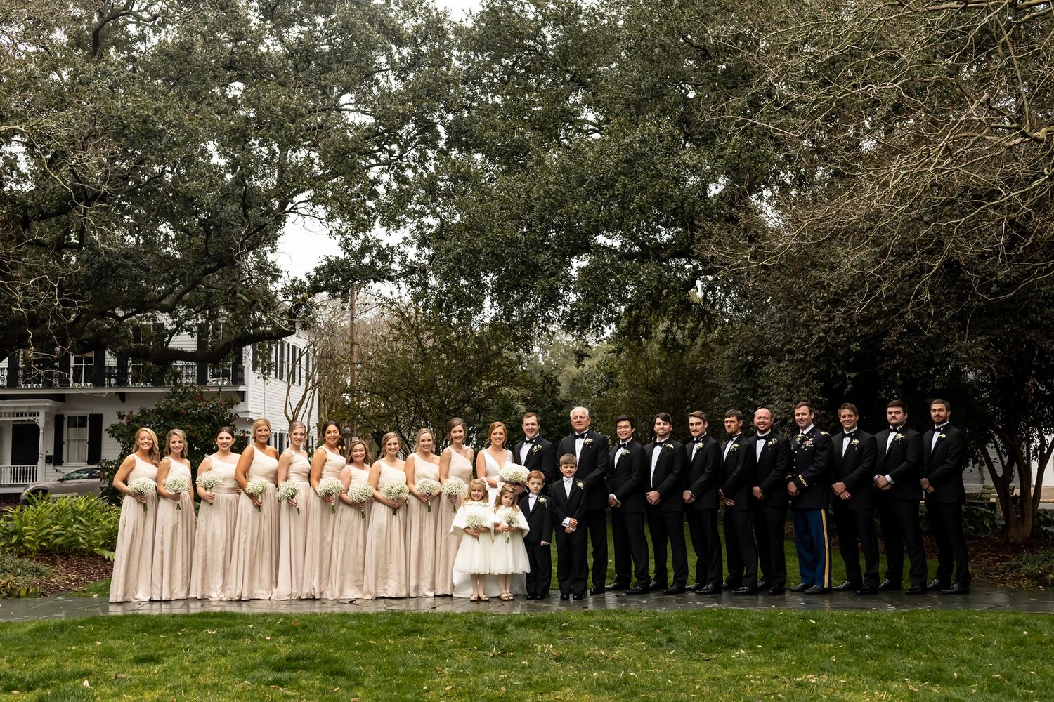 Large wedding party in formal attire lined up on green lawn with traditional black and beige color scheme under trees.