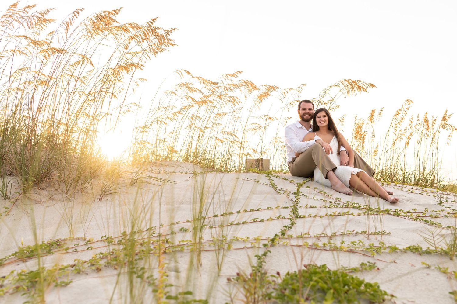 A couple sits together on a sandy beach dune with tall sea oats at sunset casting a warm golden glow.