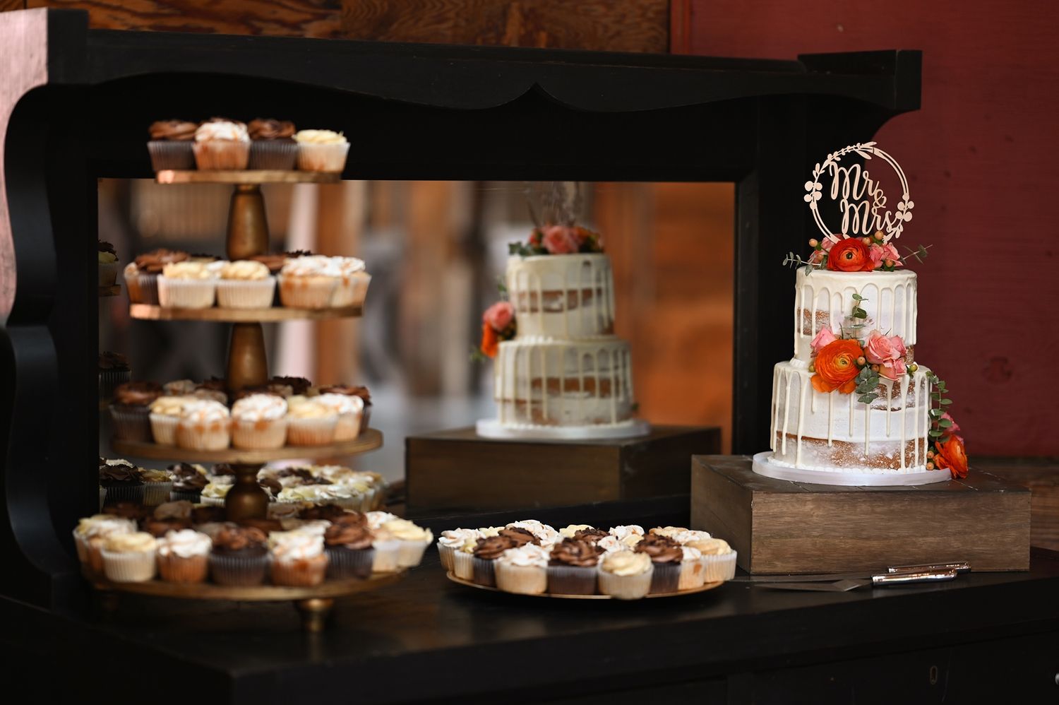 Elegant wedding dessert display featuring tiered cakes and cupcakes arranged on rustic stands against a dark background.