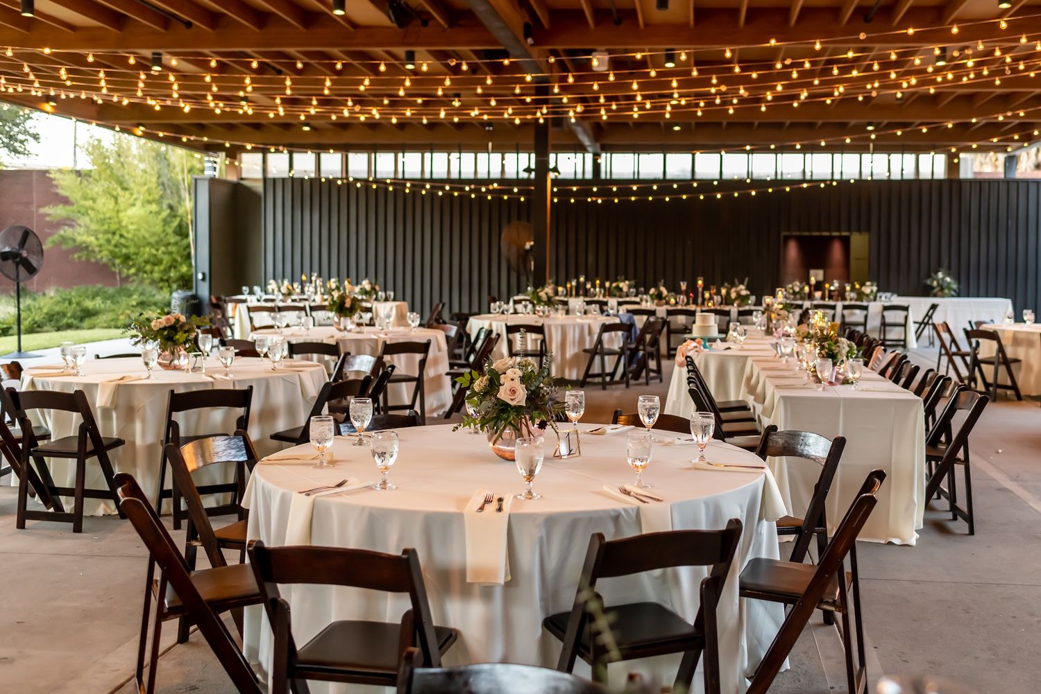 A rustic wedding reception venue with string lights overhead and round tables with white linens set for dining.
