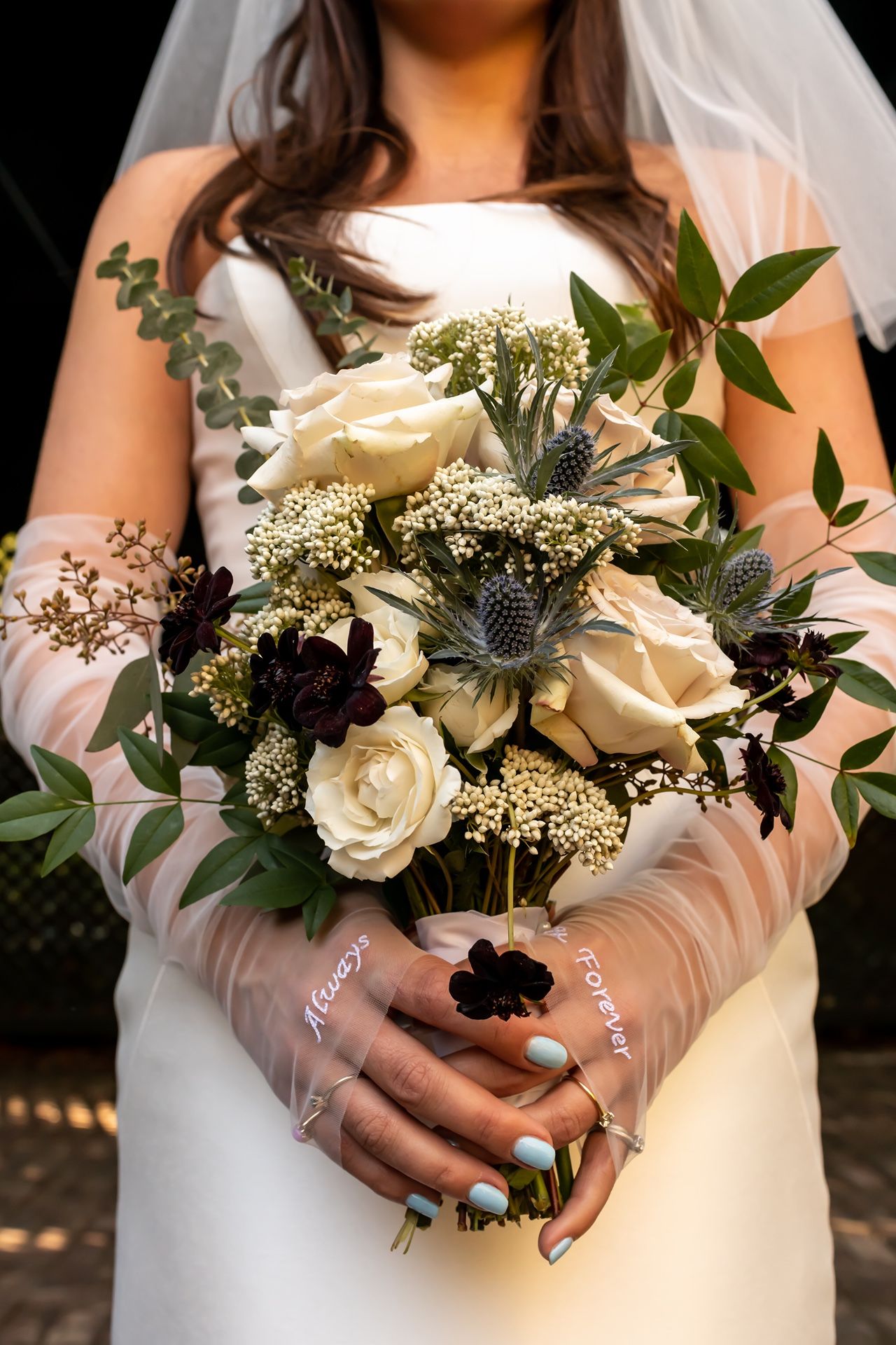 Close-up of bride holding elegant white and blue thistle wedding bouquet with roses and greenery.