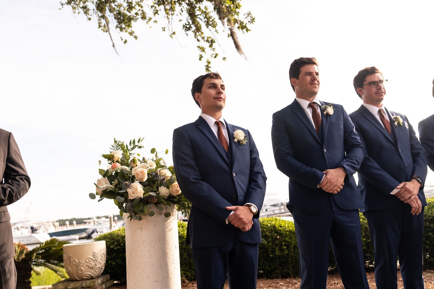 Groomsmen in navy suits stand attentively during an outdoor wedding ceremony overlooking the water.