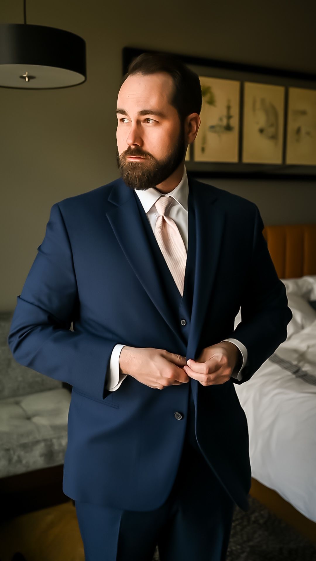 A person wearing a navy blue three-piece suit and cream tie adjusts his jacket buttons in a dimly lit hotel room.