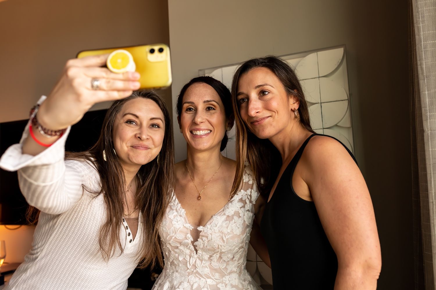 Three friends take a selfie together at a party, with one wearing an elegant lace white dress.