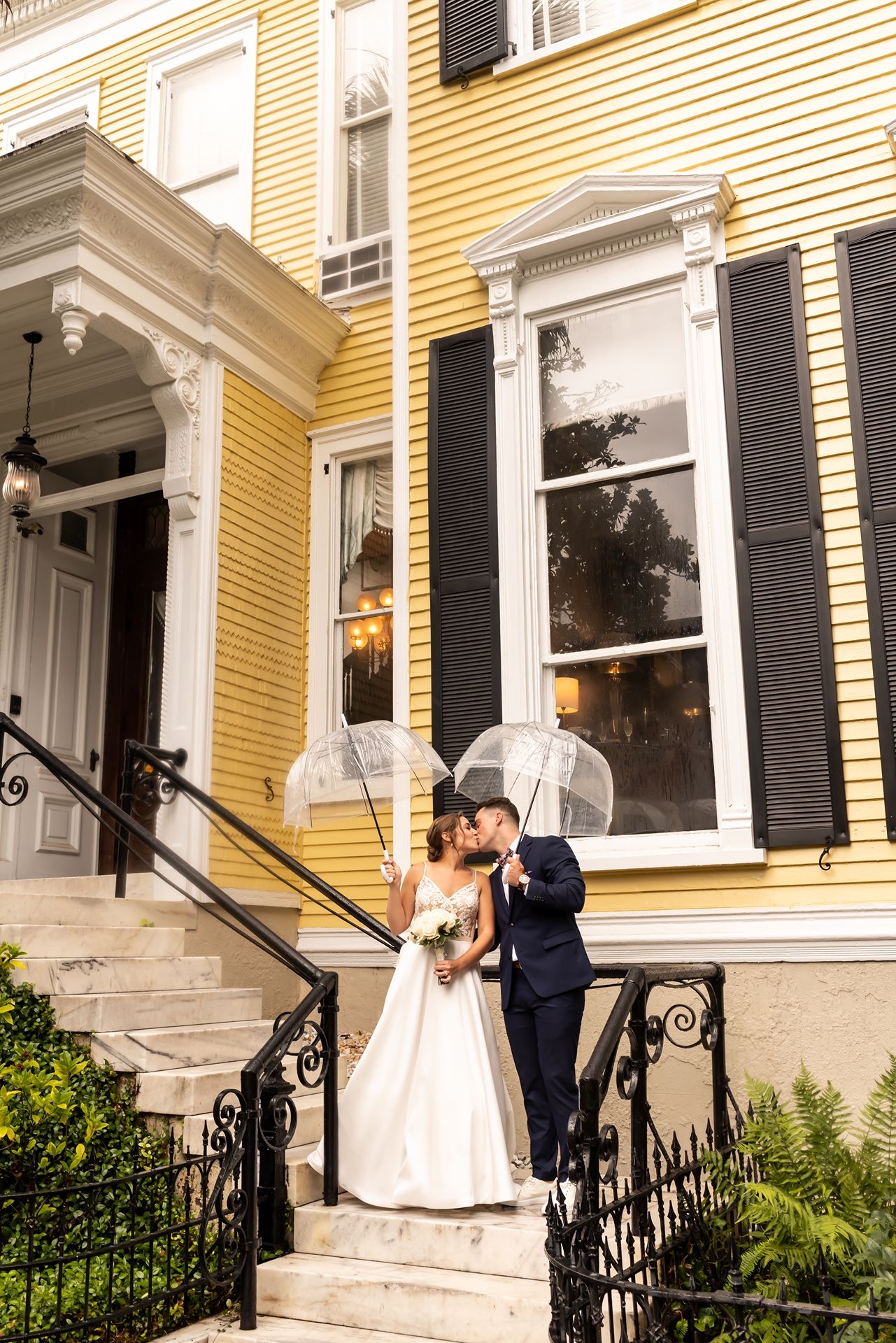A house with yellow siding and white trim with an elegant entrance stairway and black iron railings on a bright day.