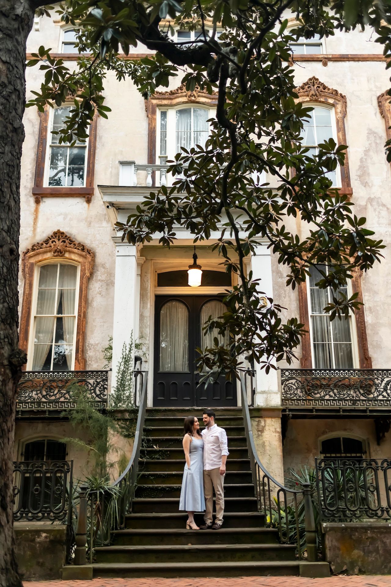 Historic brownstone building with ornate iron railings and steps leading to multiple entrances framed by magnolia trees.