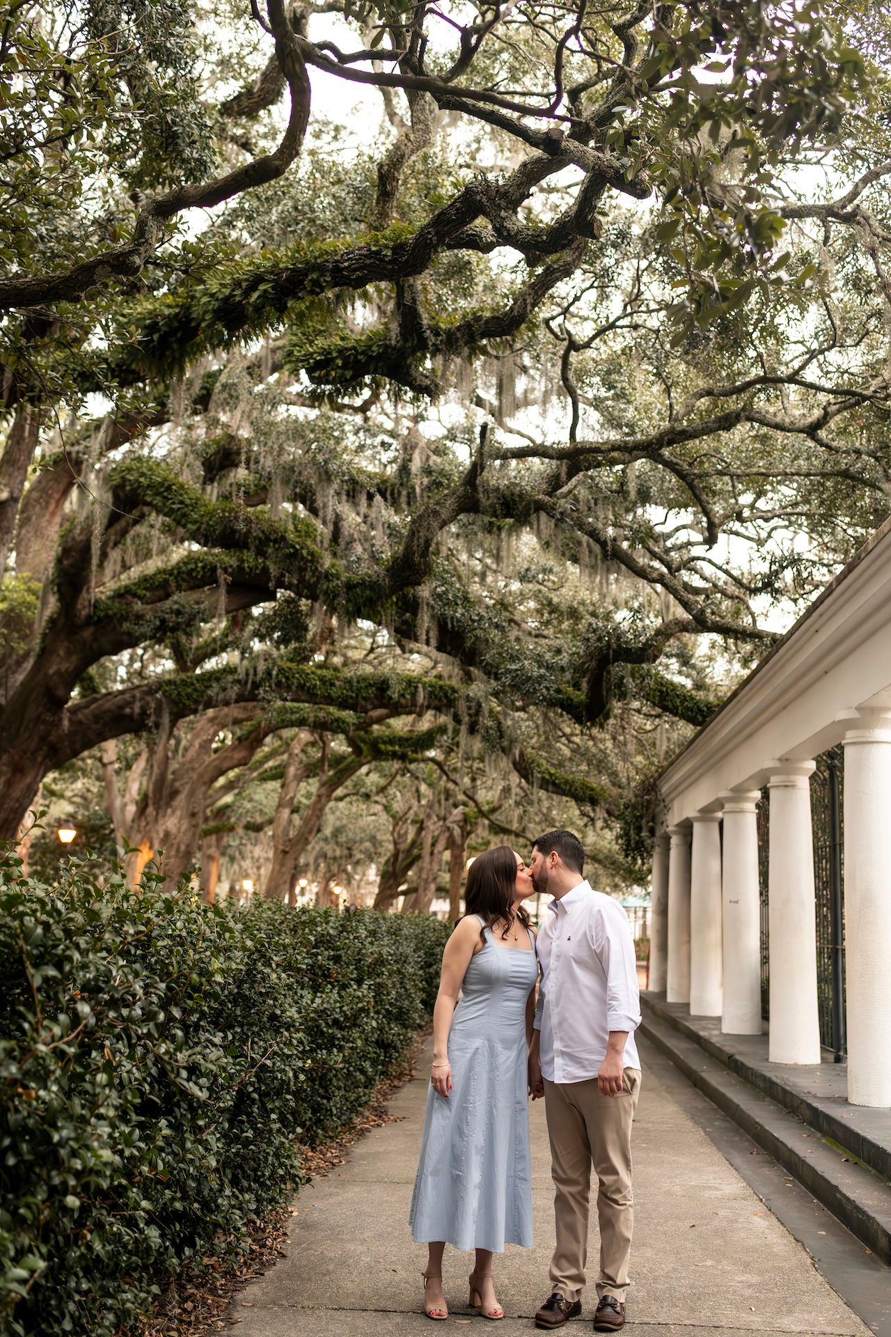 A couple stands close together on a path beneath moss-draped oak trees and white columns in the Southern United States.