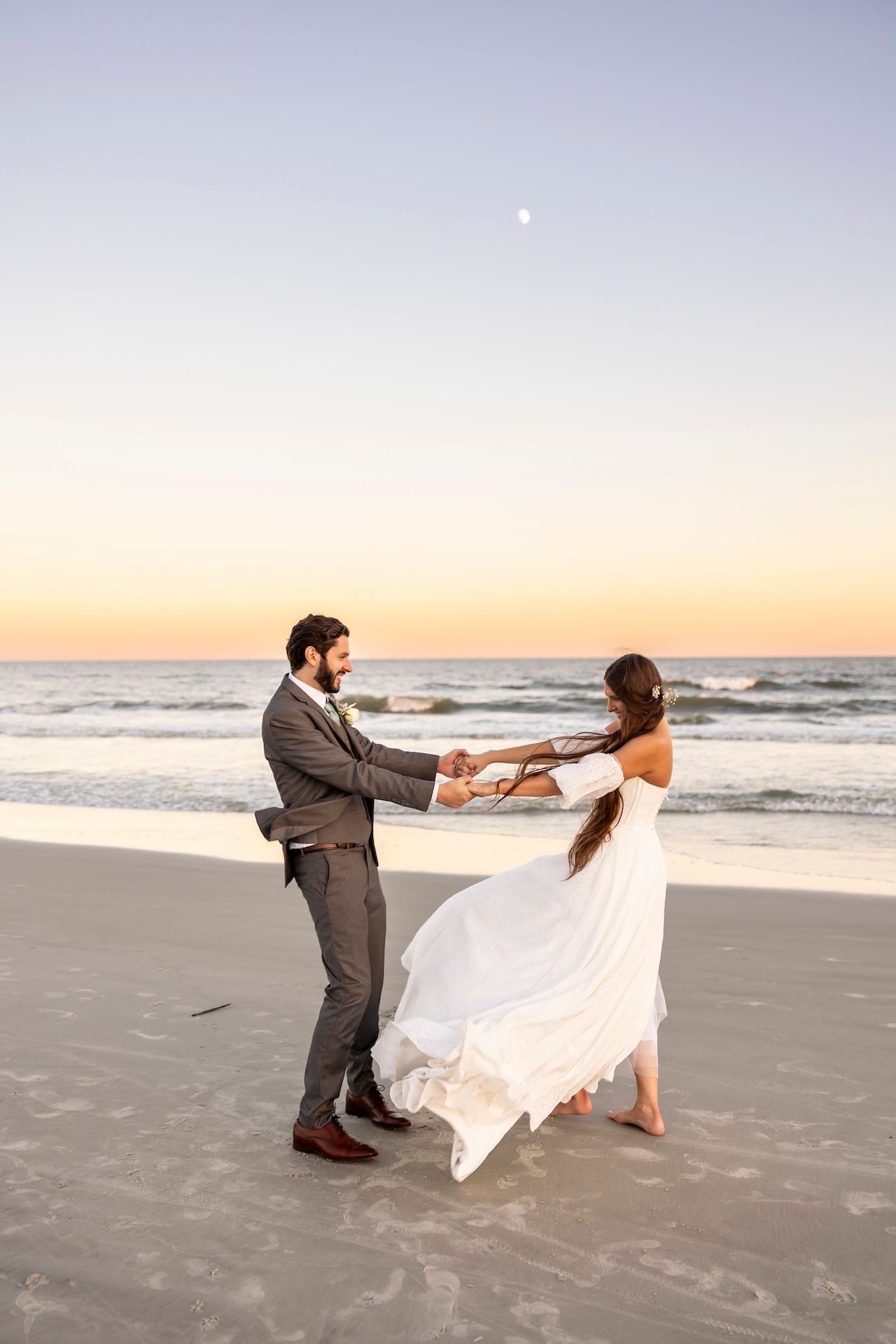 Newlyweds dance together at sunset on a beach with white dress flowing in ocean breeze.