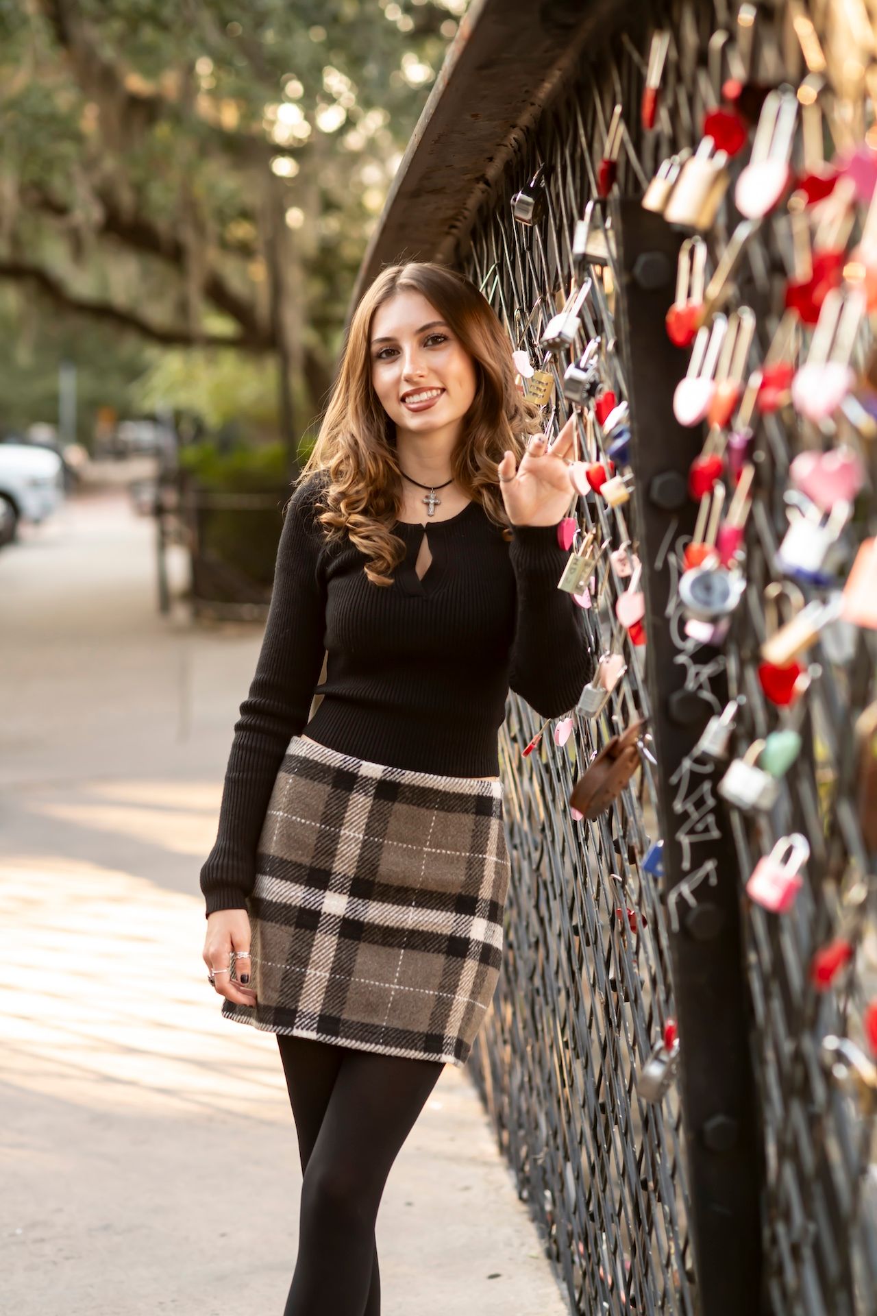 Woman in black sweater and plaid skirt poses next to wall decorated with many hanging locks on city street.