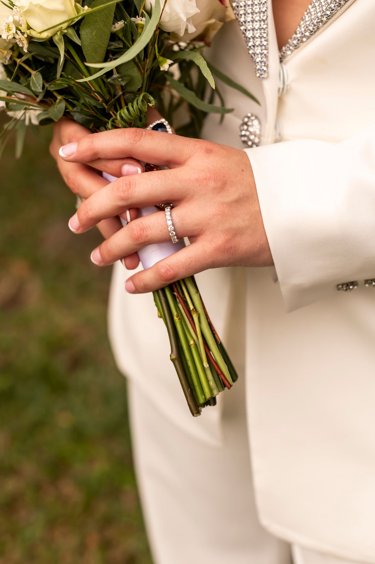 Hand wearing a sparkling engagement ring holds a white rose bouquet wrapped in elegant white silk ribbon.