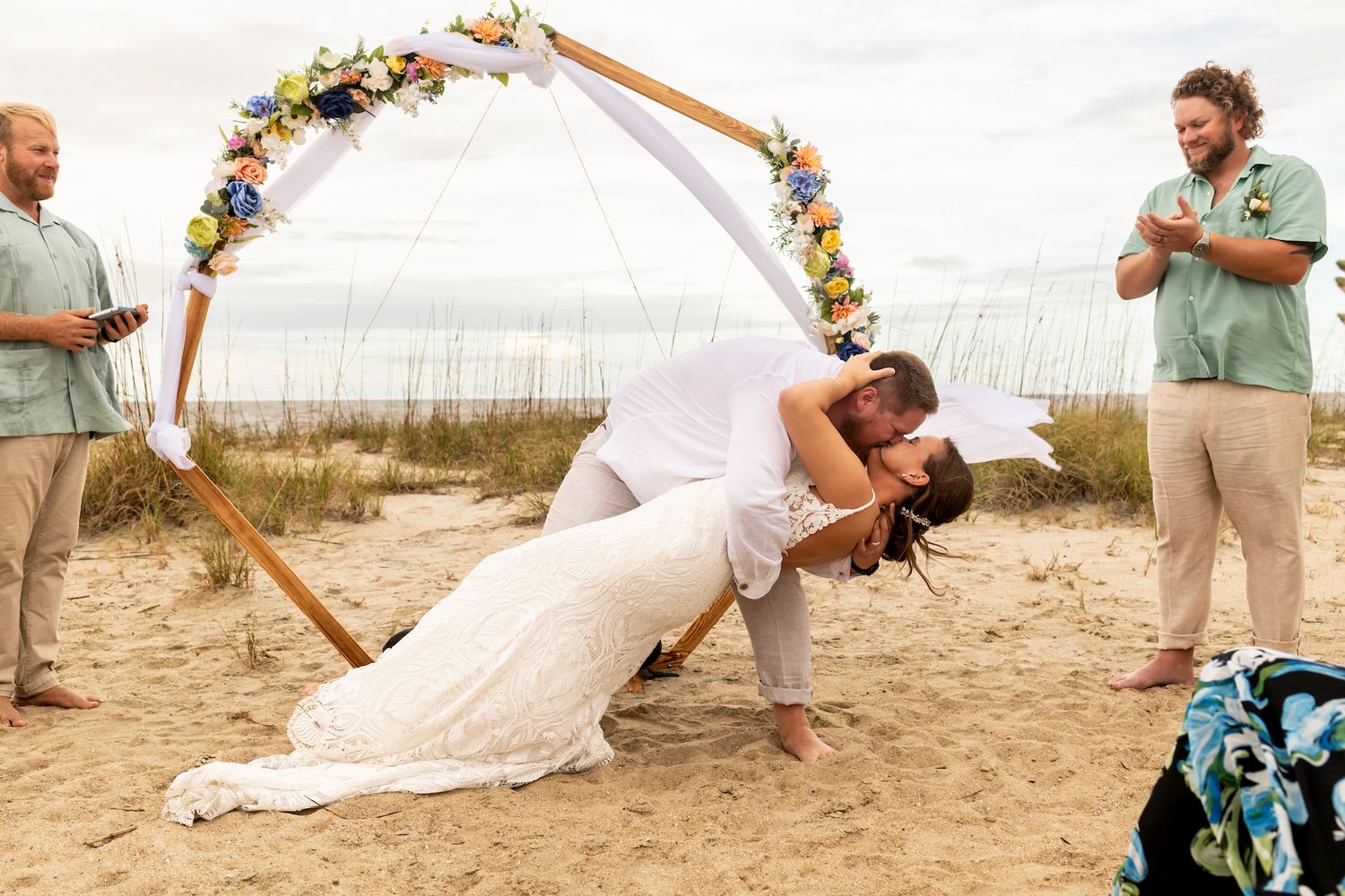 A romantic dip kiss under a floral arch on the beach during a destination wedding ceremony.
