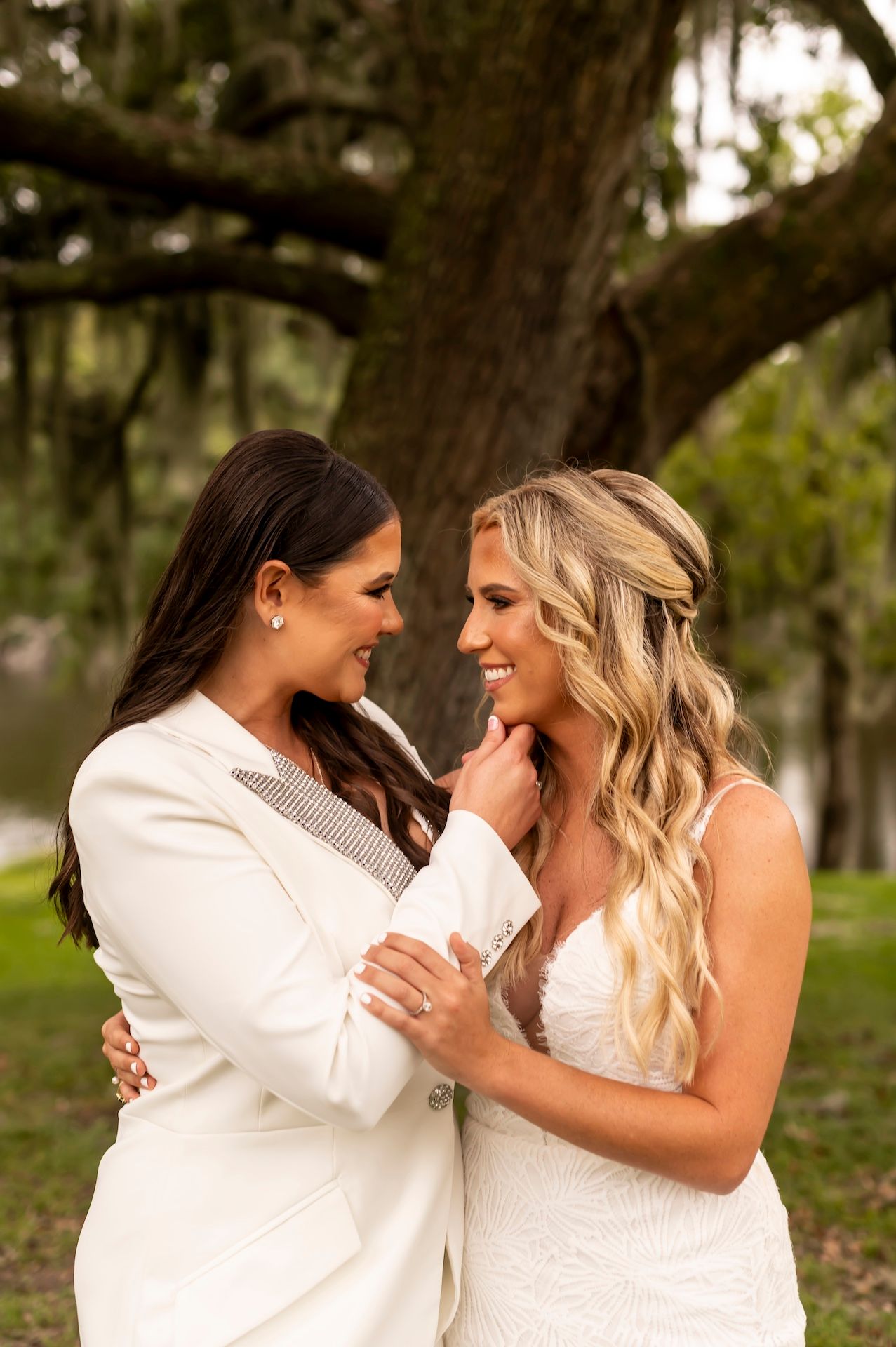 Two people in white wedding attire share an intimate embrace while smiling in a garden setting with a large tree behind them.