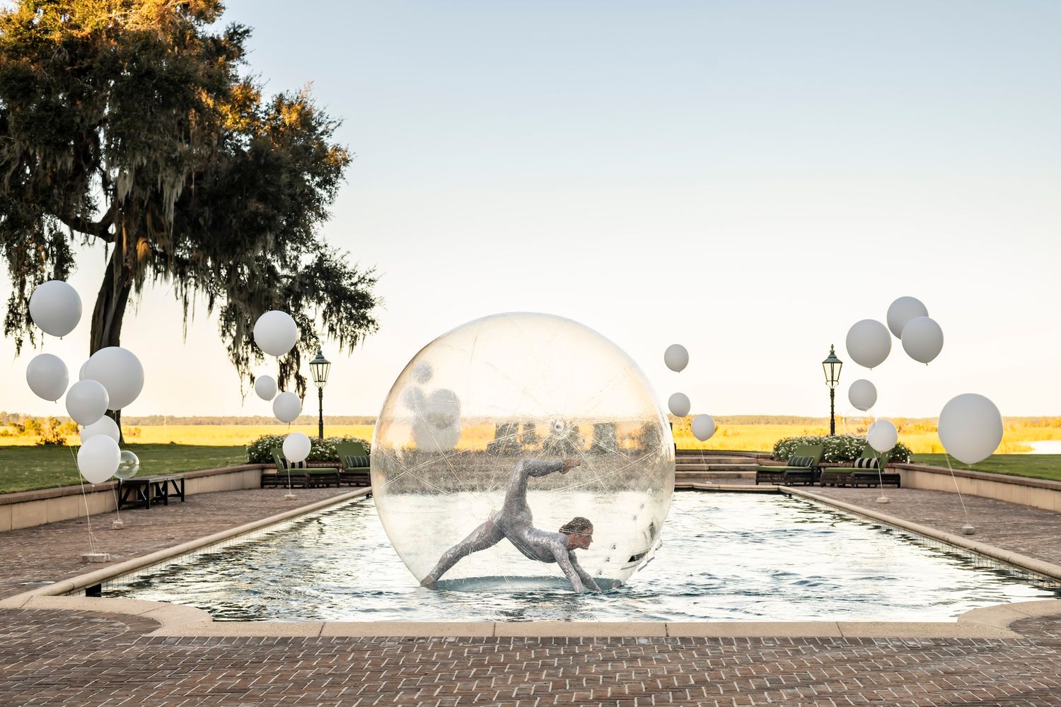 A performer in a large transparent bubble dances on a reflecting pool surrounded by white balloon decorations at sunset.