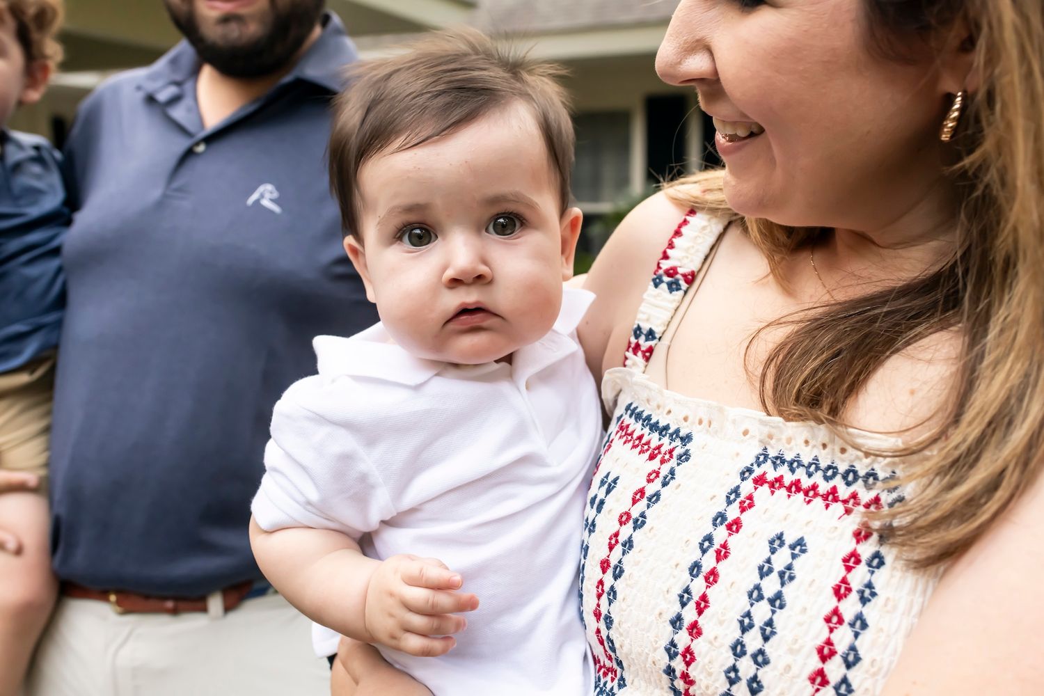 A baby wearing white clothing being held outdoors on a sunny day.