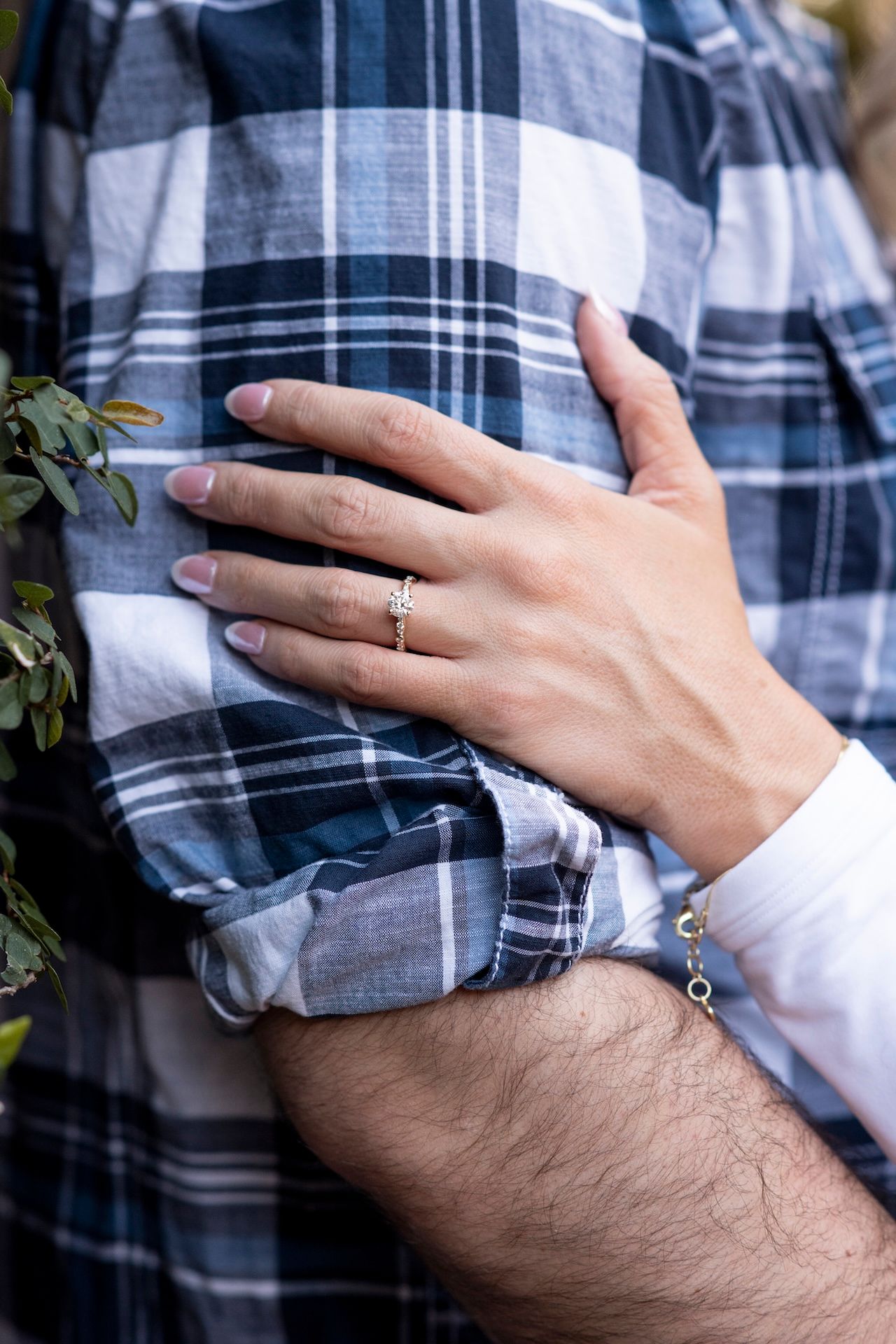 Close-up of hands embracing against plaid shirt showing engagement ring and bracelet with greenery in background.