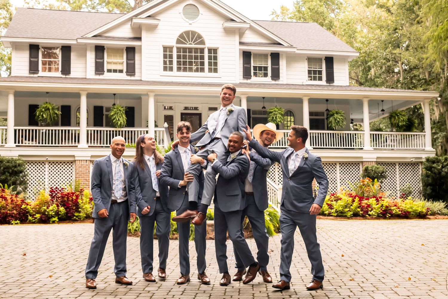 Joyful groomsmen in gray suits lift and carry someone while posing playfully on a brick courtyard.