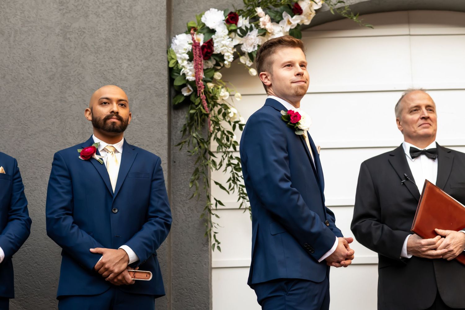 Wedding ceremony floral arch adorned with pink and white roses against a white wall with elegantly dressed attendees.