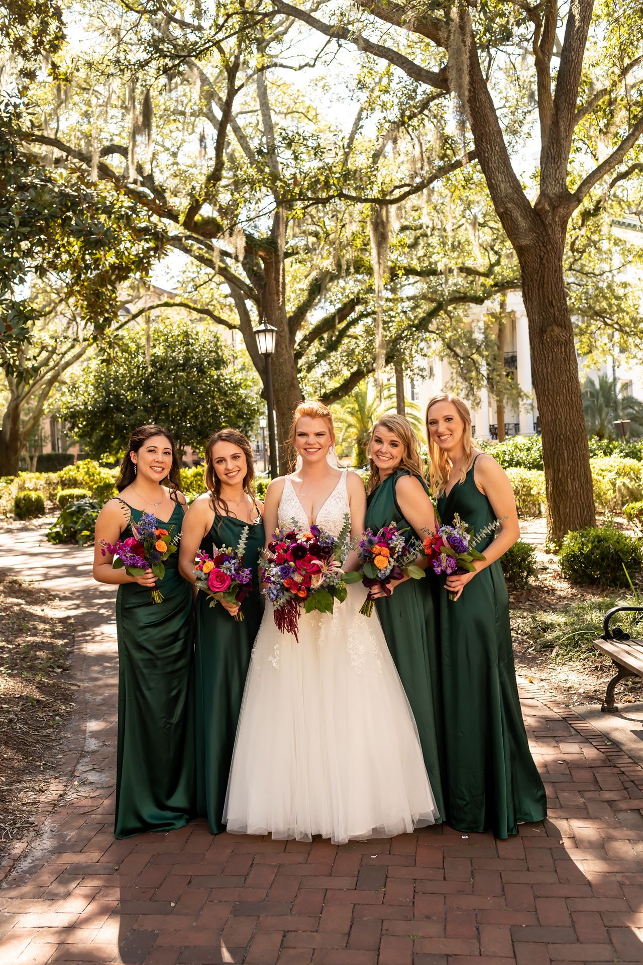 Wedding party poses on brick path under Spanish moss draped trees wearing emerald green gowns and holding vibrant bouquets.