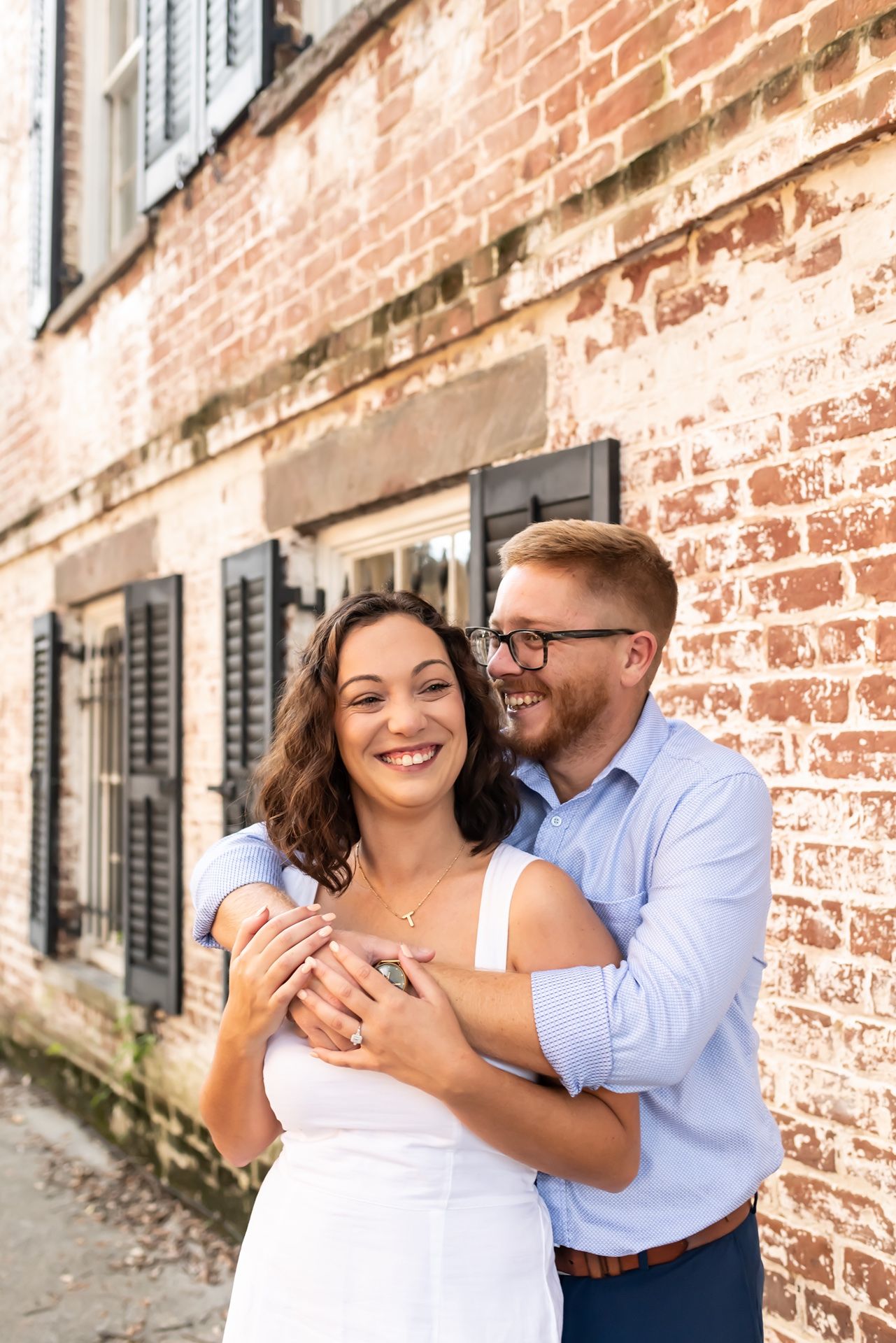 Happy couple embraces and laughs together against weathered brick wall on sunny day.