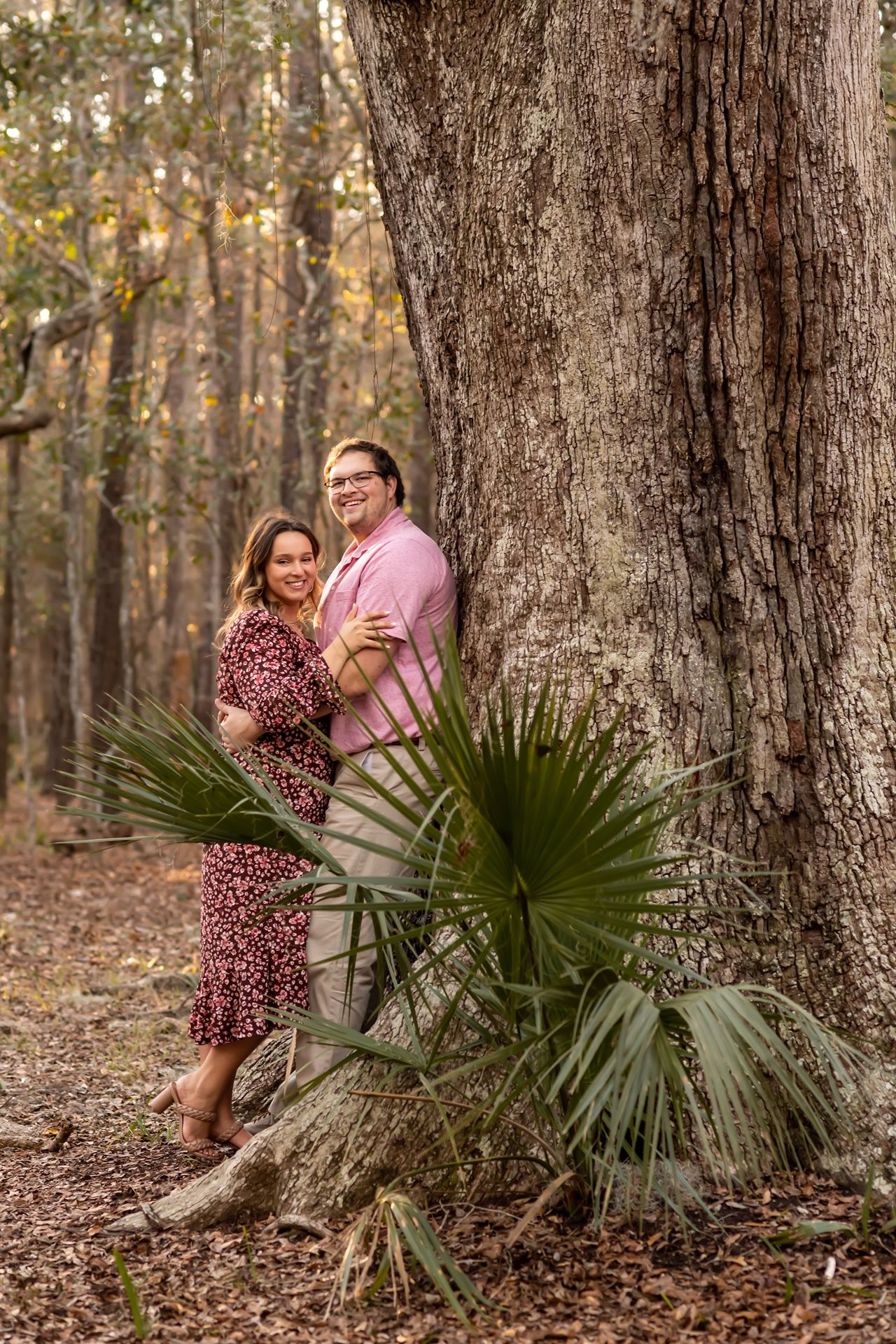 Couple stands together against large tree trunk in autumn forest with palmetto plants at base.