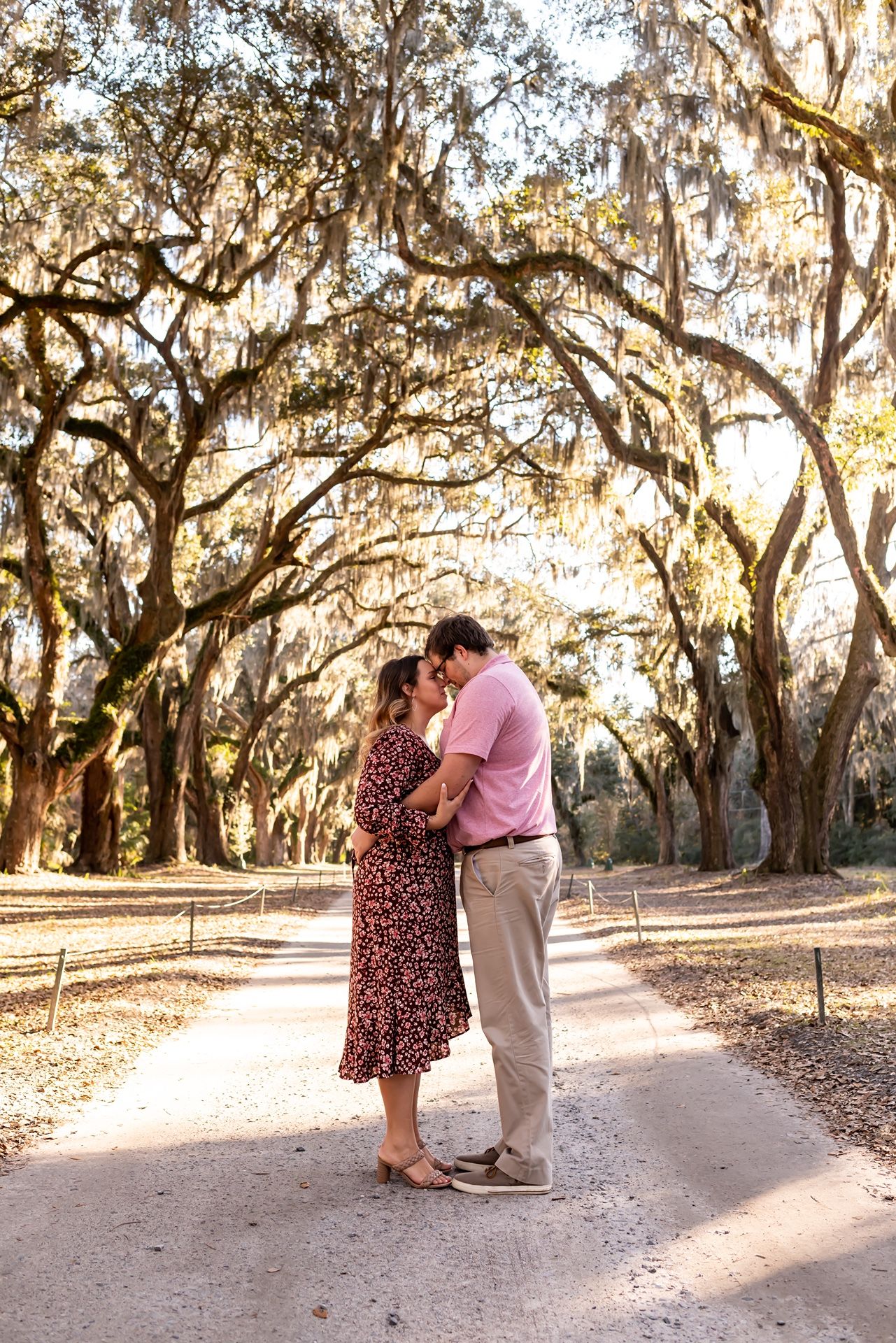 A couple shares a romantic moment on a path lined with sunlit Spanish moss-draped oak trees.