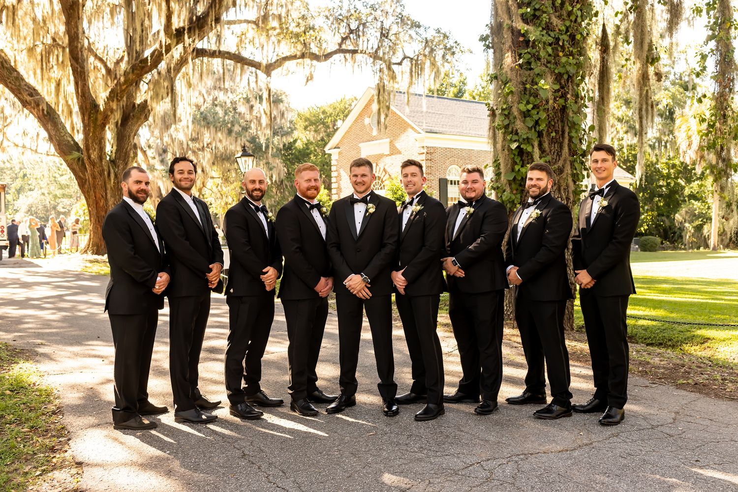 A formal group in black tuxedos poses together on a tree-lined path at an outdoor venue.