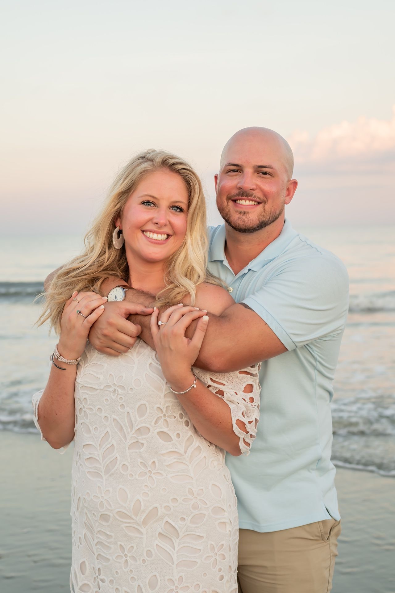 A couple shares a romantic moment at sunset on the beach, wearing white and light blue casual attire.