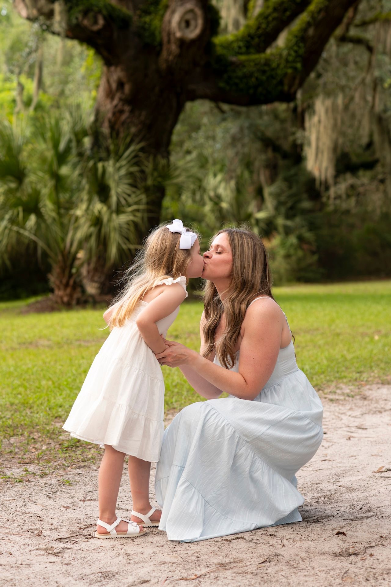 A mother in a blue dress kisses her young daughter in a white dress outdoors near a mossy oak tree.