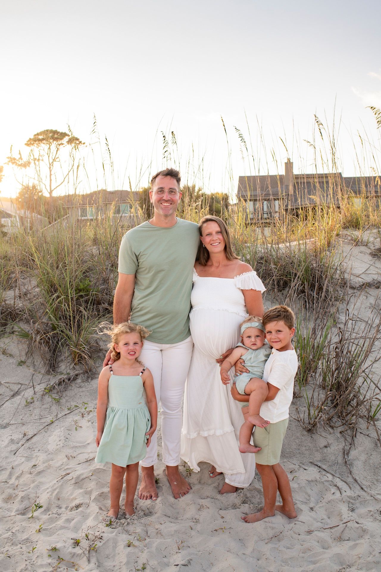 Family of five poses on sandy beach dunes at sunset, parents with three young children, woman in white maternity dress.