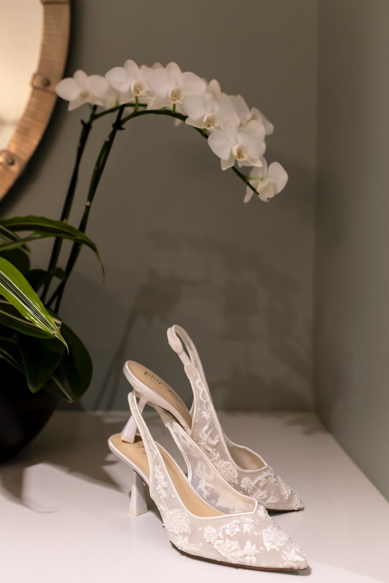 White lace slingback bridal heels displayed beside delicate white orchids on a light surface create an elegant wedding scene.