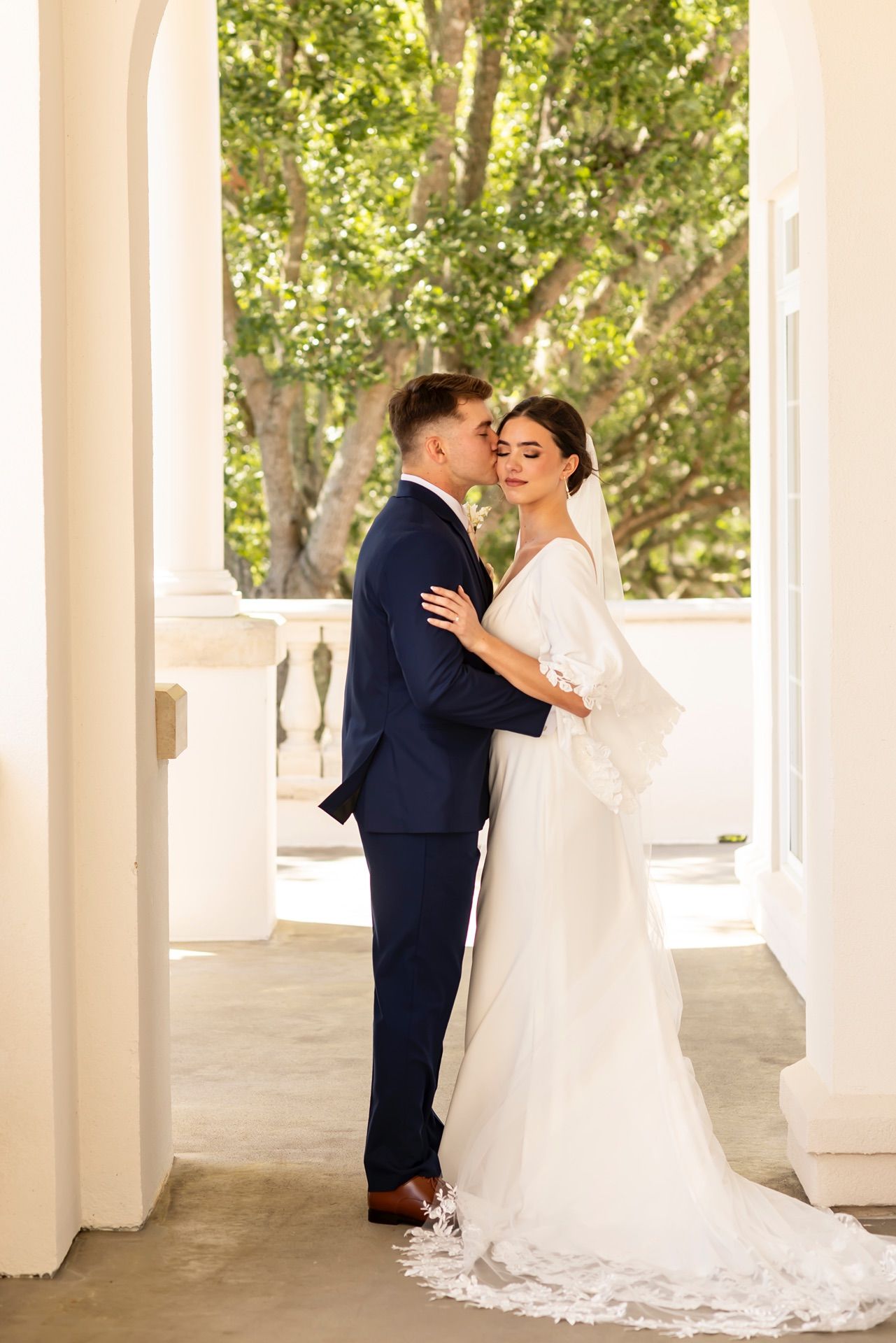 A romantic wedding scene under a covered walkway with white columns and green trees visible in the background.