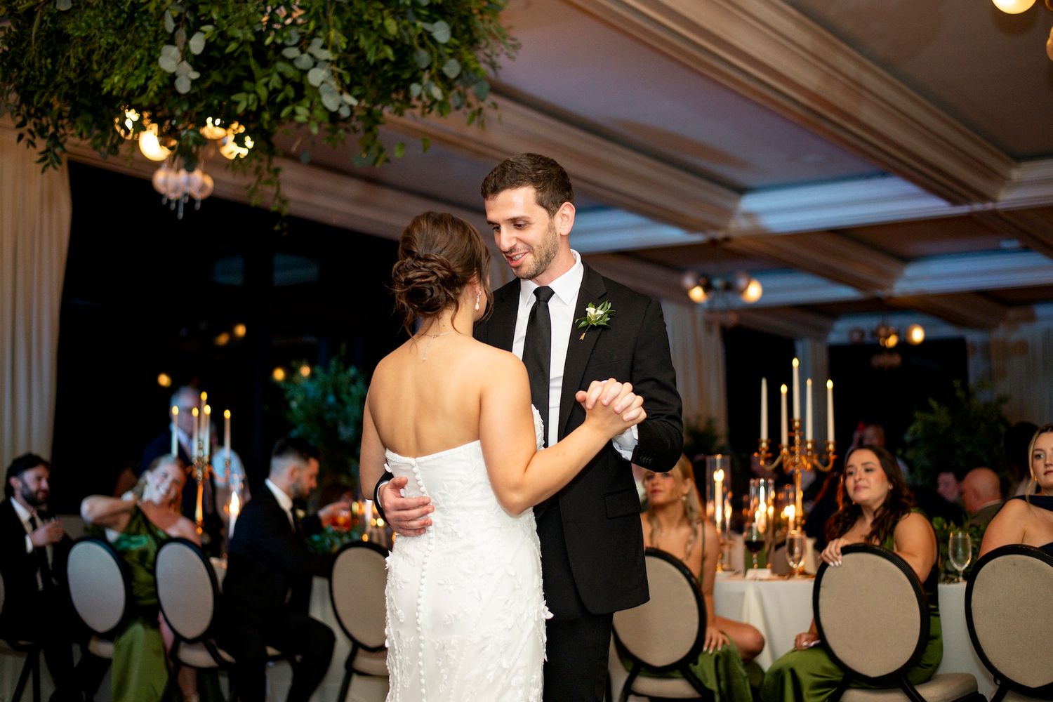 A couple shares their first dance at an elegant wedding reception with chandeliers and greenery decorations.