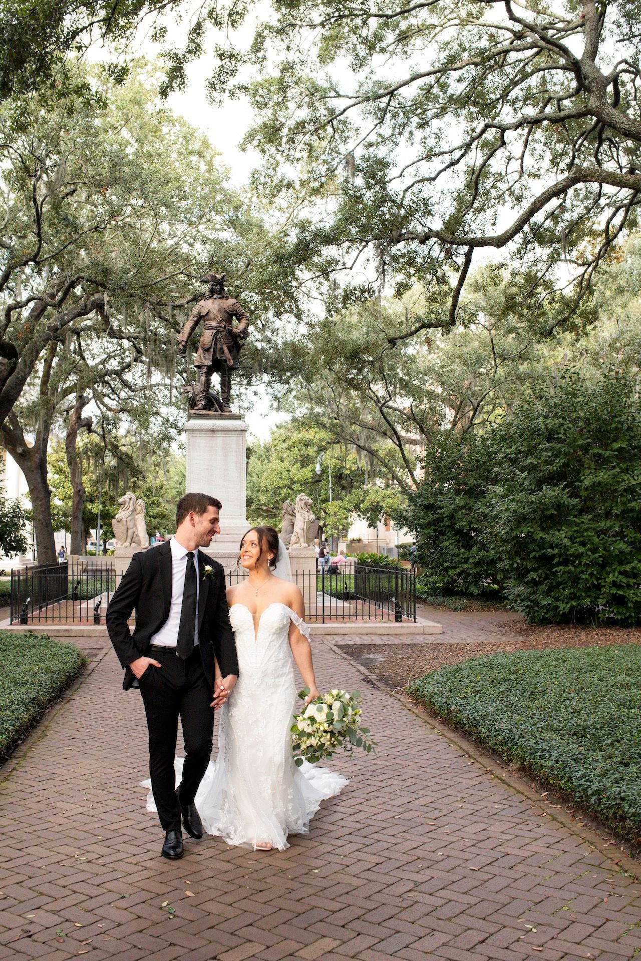 Newlyweds share intimate moments while walking along a brick pathway surrounded by lush greenery and oak trees.