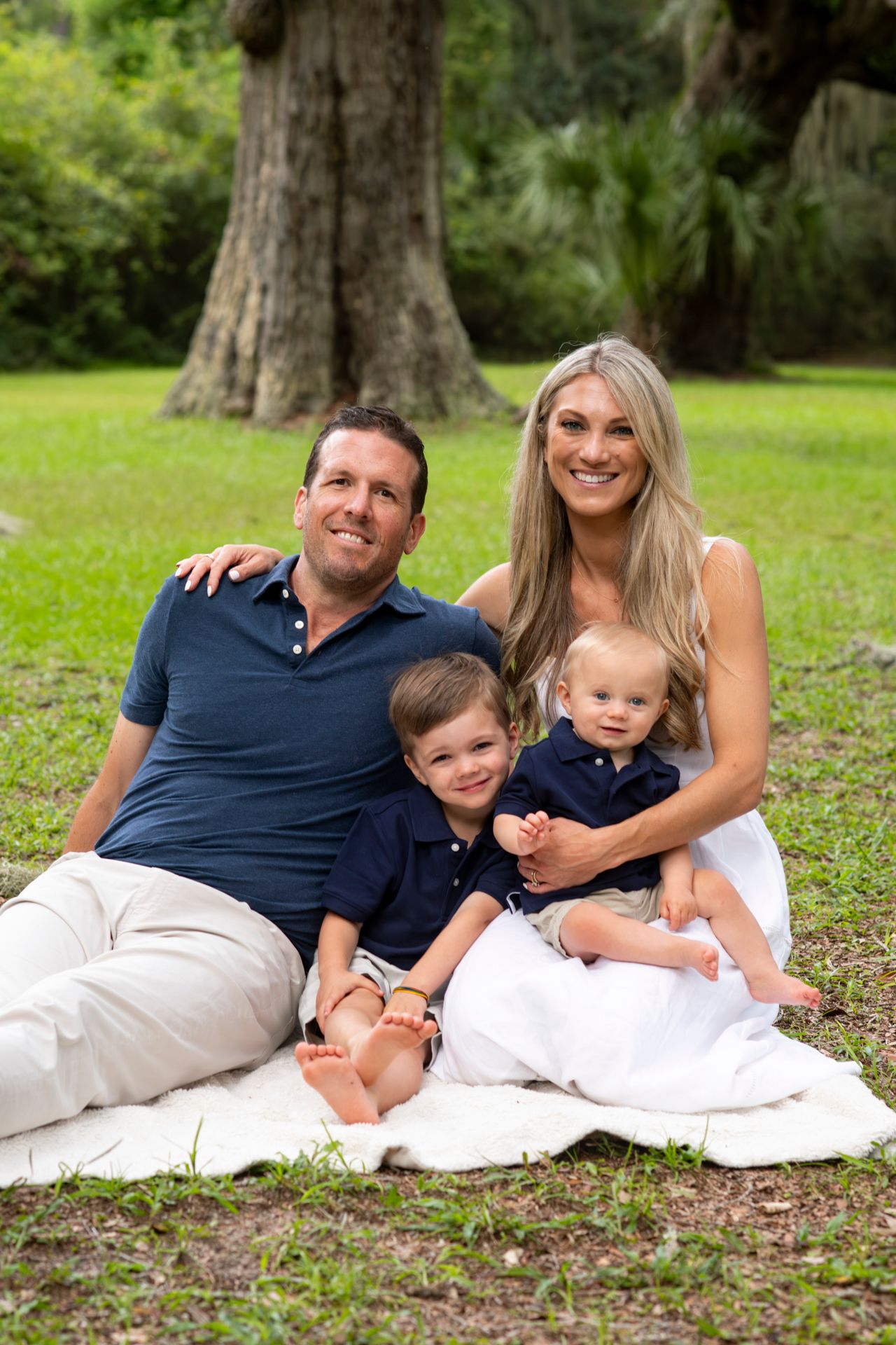 A family sits together on a white blanket for an outdoor portrait session under large oak trees.