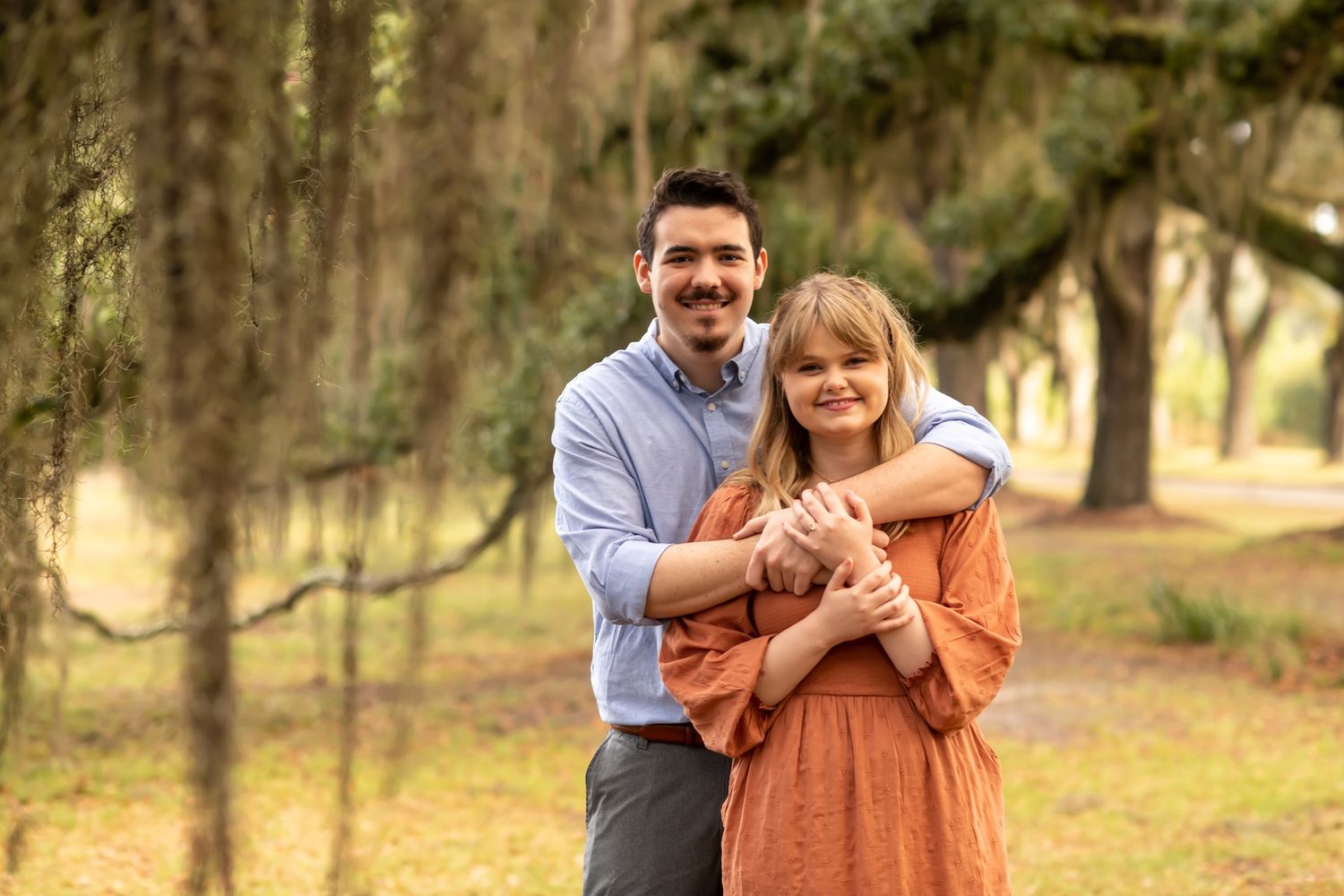 A couple embraces in a romantic outdoor photoshoot at a wooded park during golden hour.