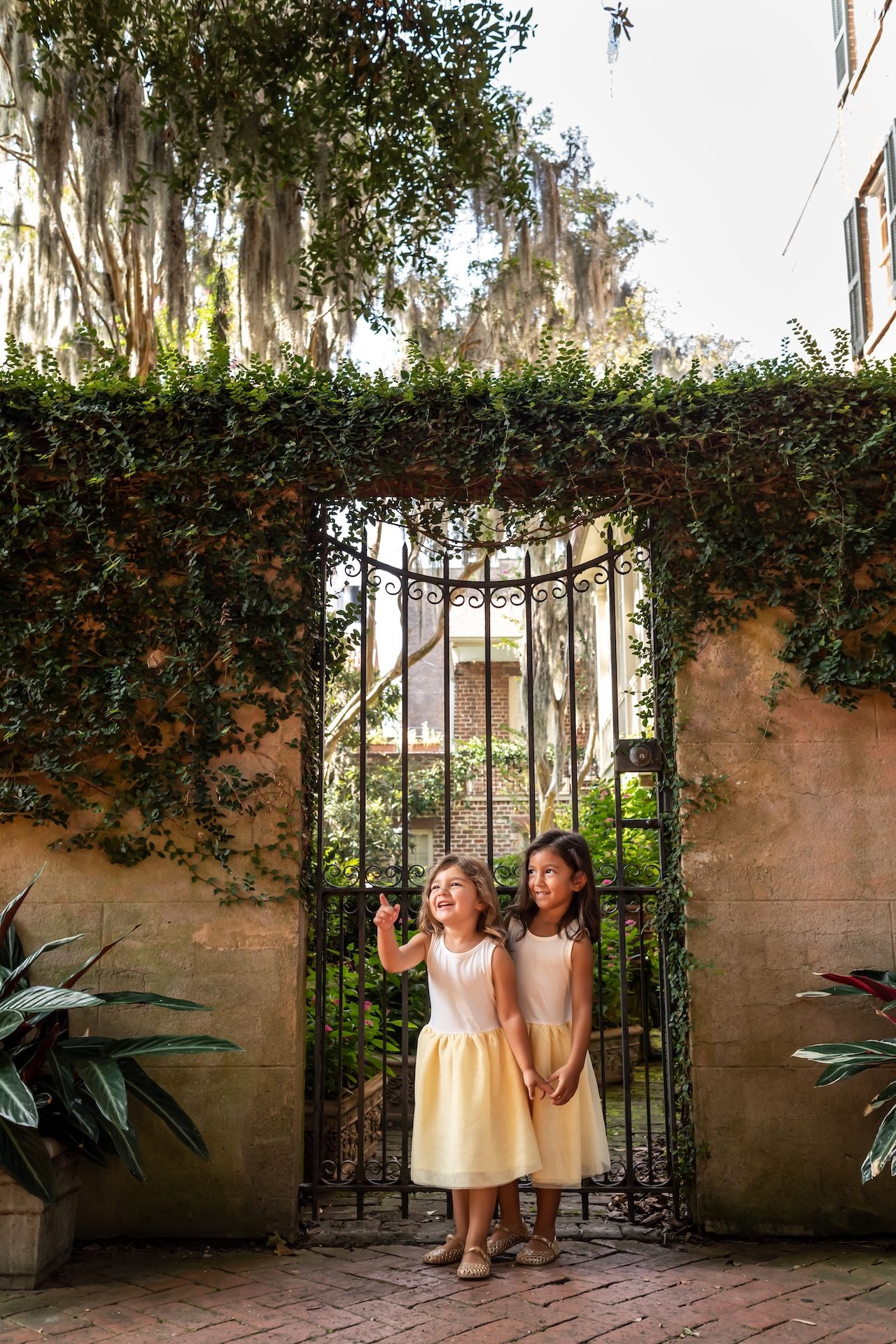 Two girls in yellow dresses stand near an ornate garden gate for a family portrait on Jones Street in Savannah.