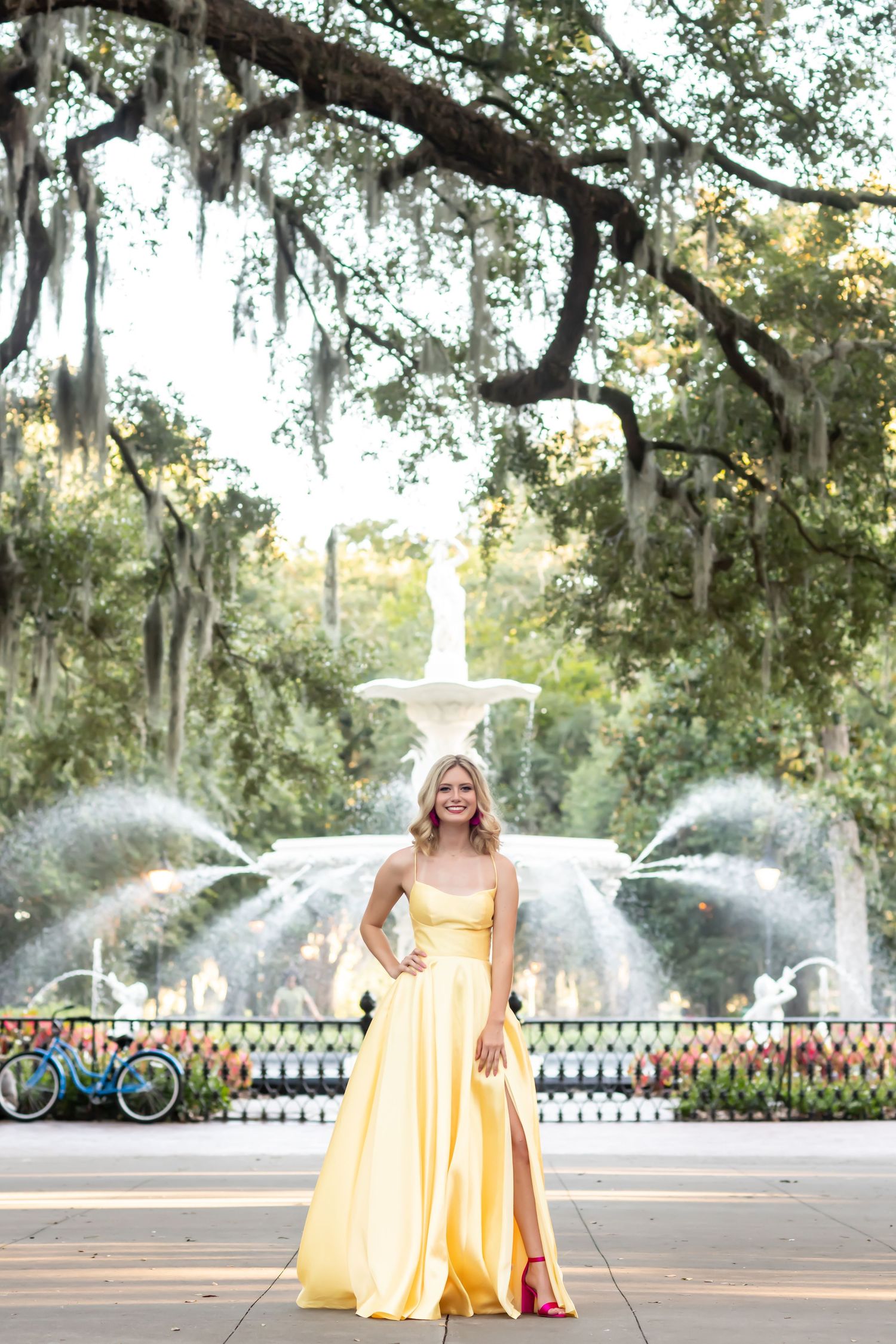 Senior graduate in yellow formal dress poses by fountain with Spanish moss draped trees in background.