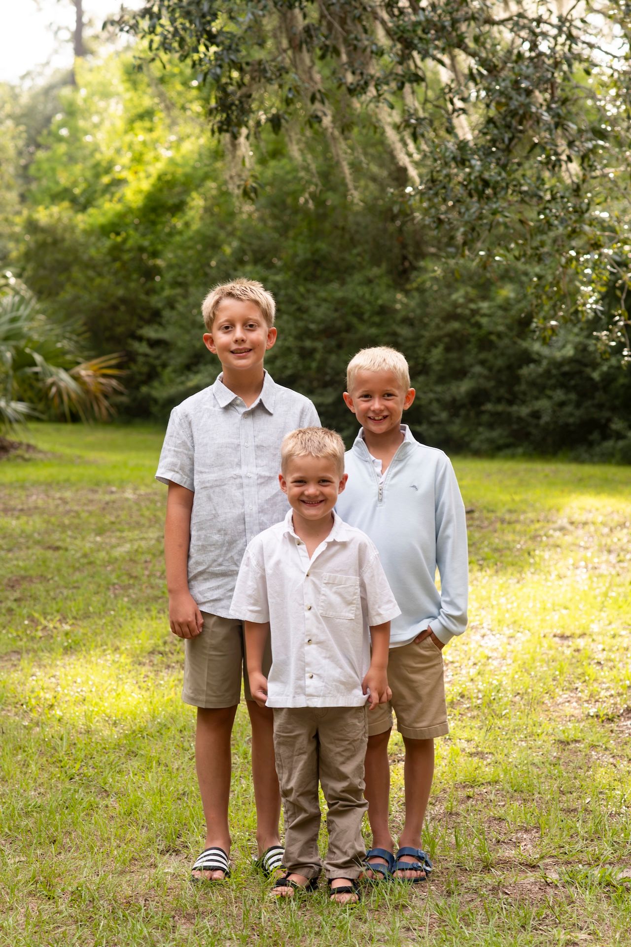 Three young blonde boys posing outdoors in casual button-up shirts and khaki shorts, smiling in a lush green park setting.
