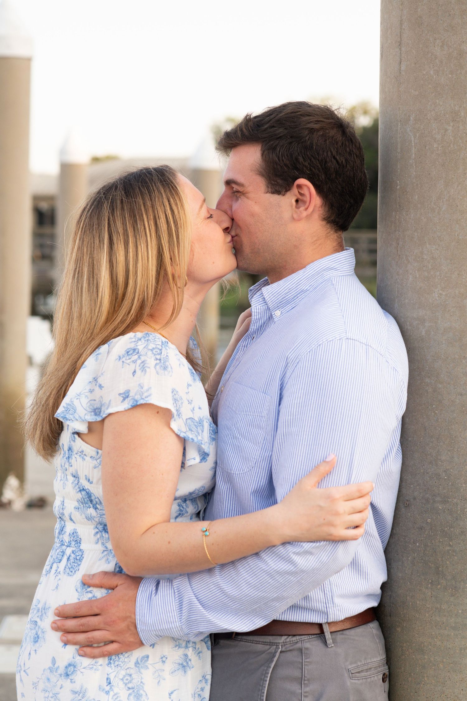 A romantic couple shares a tender kiss while embracing near a stone column, she in a floral dress and he in a blue shirt.
