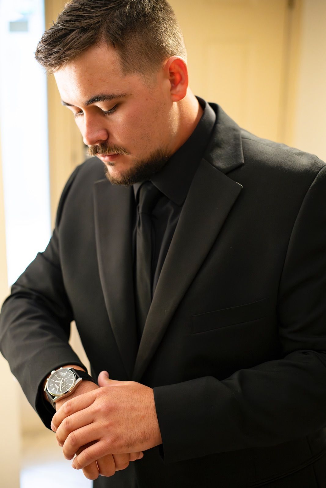 A man in a black suit adjusts his watch while getting ready for a formal event.