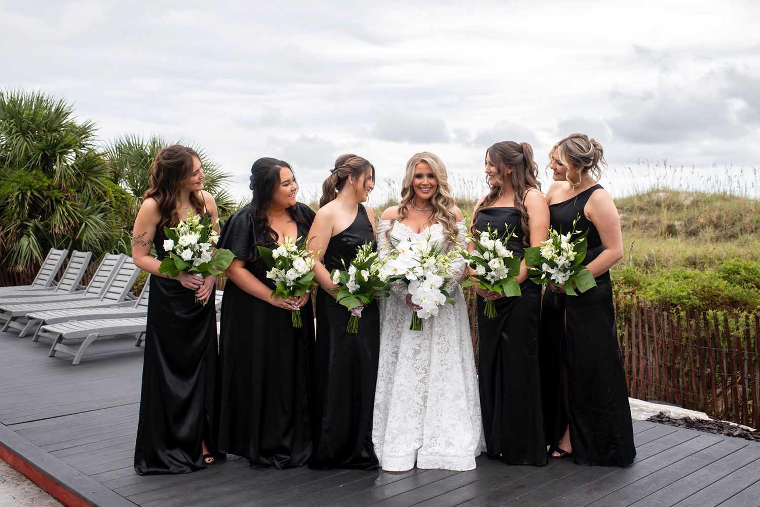 A wedding party in black and white attire poses outdoors with bouquets against a natural landscape backdrop.