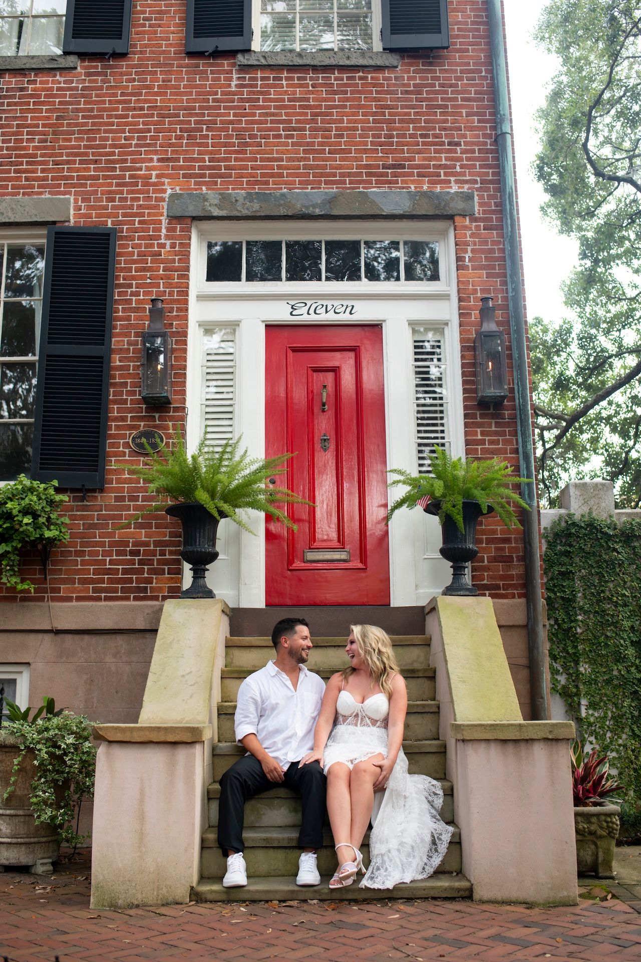 Couple sitting together on brick steps outside a historic red brick townhouse with a bright red door and potted palm plants.