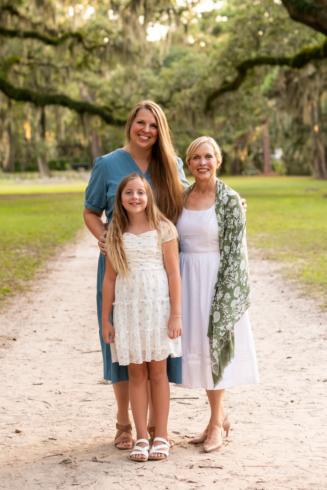 Three generations of women pose on a tree-lined path, wearing white and teal dresses in a lush outdoor setting.
