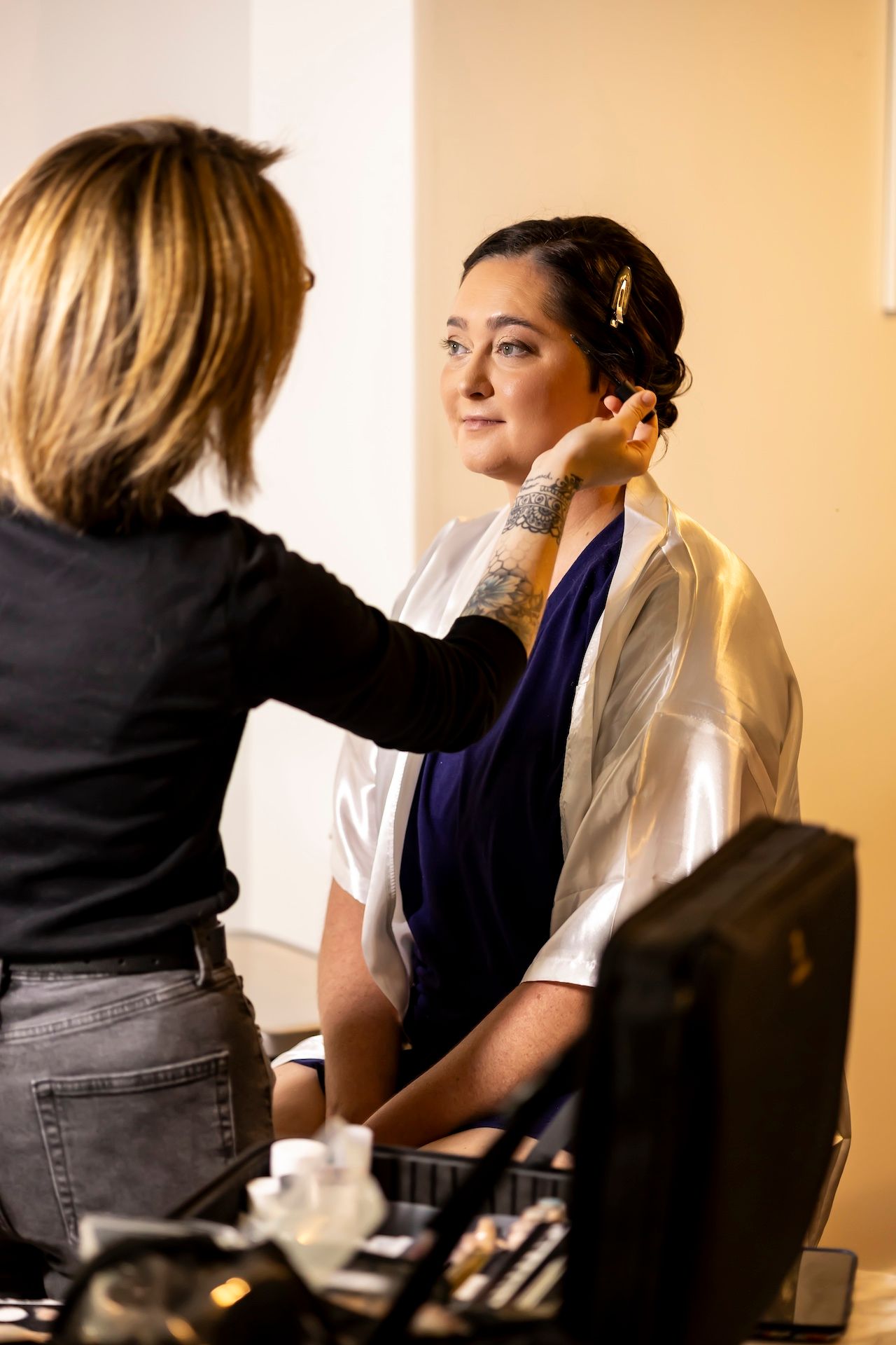 Makeup artist applies cosmetics on model in white shirt during professional beauty session with tools and products nearby.