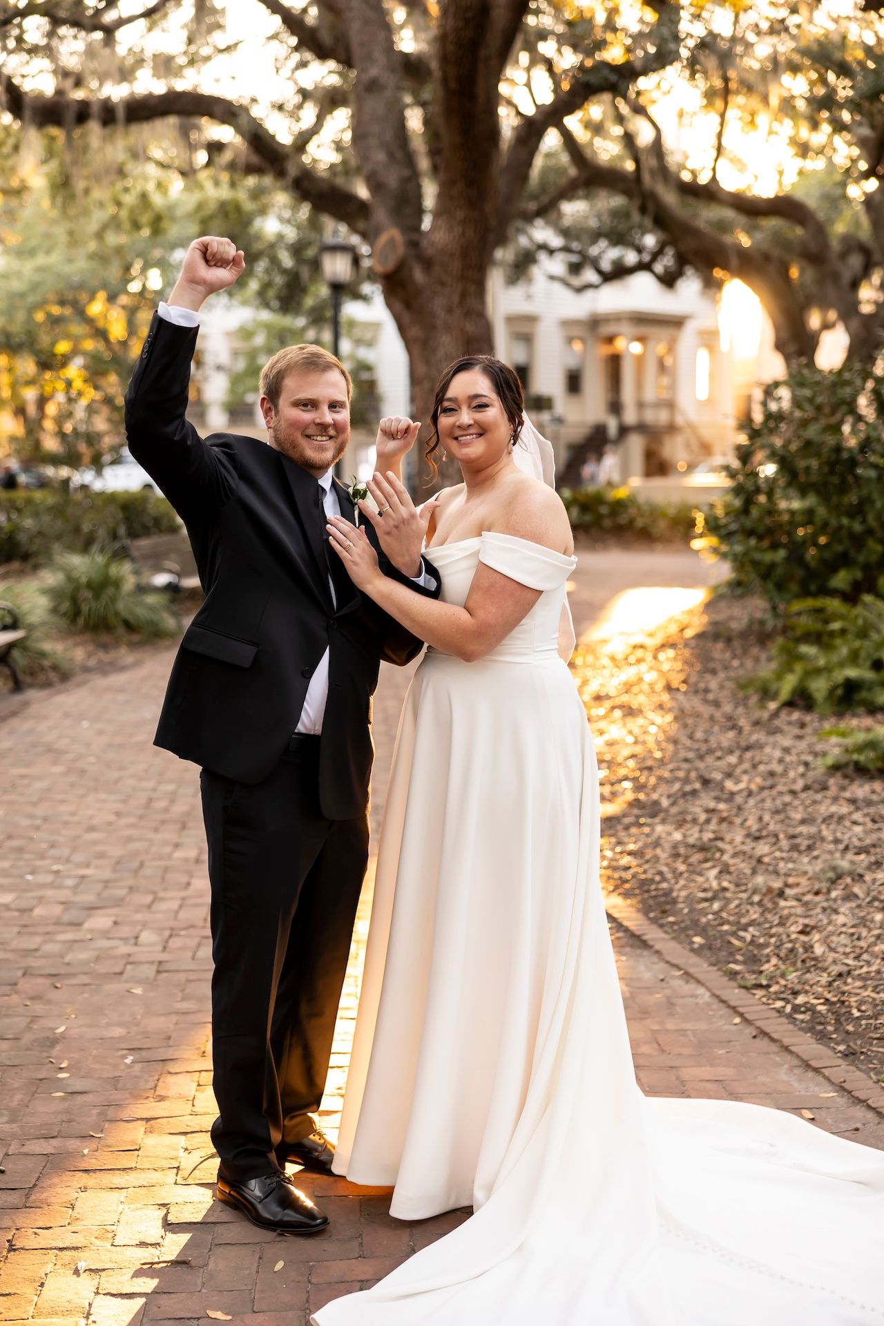 Newlyweds celebrate joyfully on a brick pathway beneath oak trees at sunset during an outdoor southern wedding.
