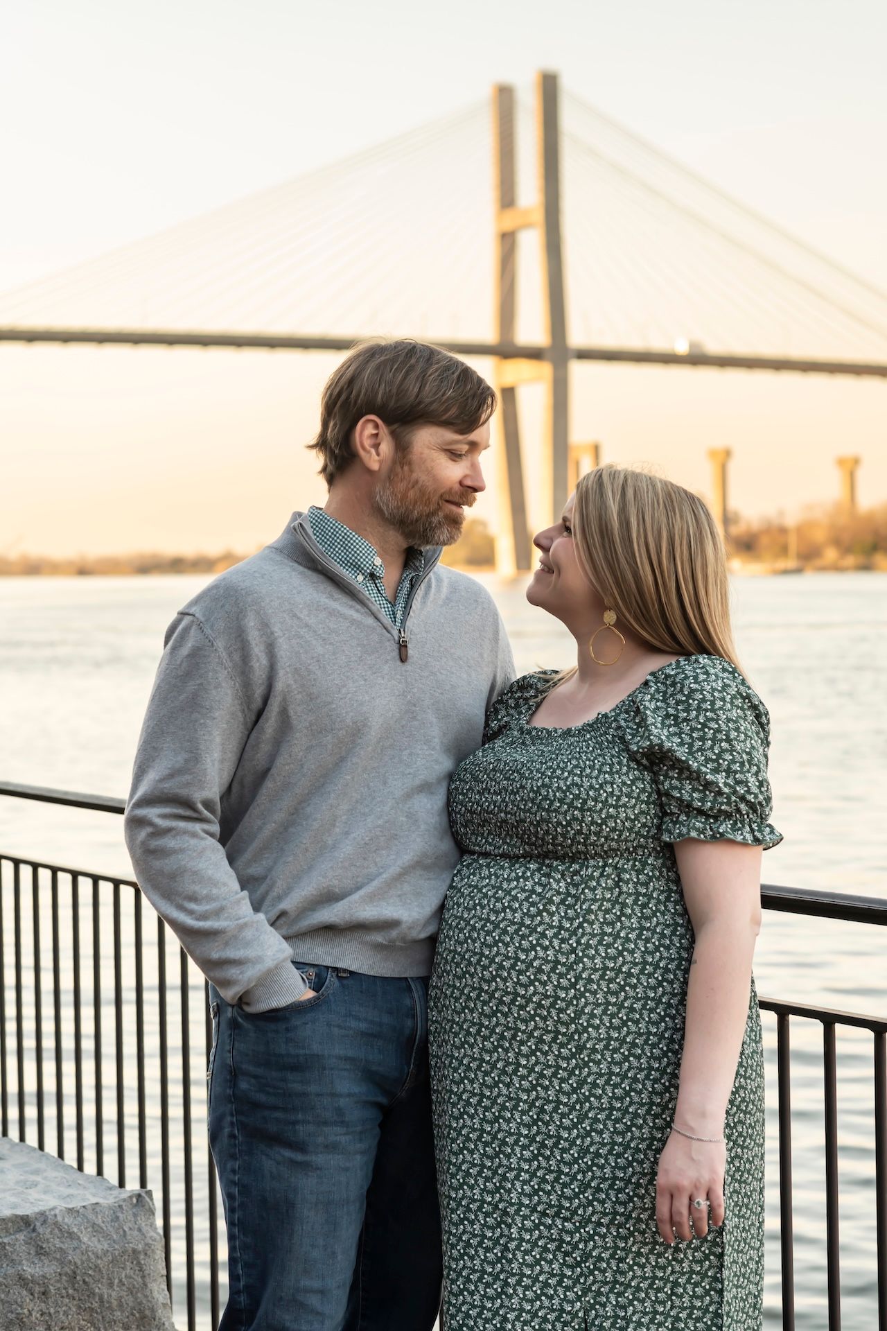 A couple stands lovingly by a waterfront railing at sunset with a bridge visible in the background.