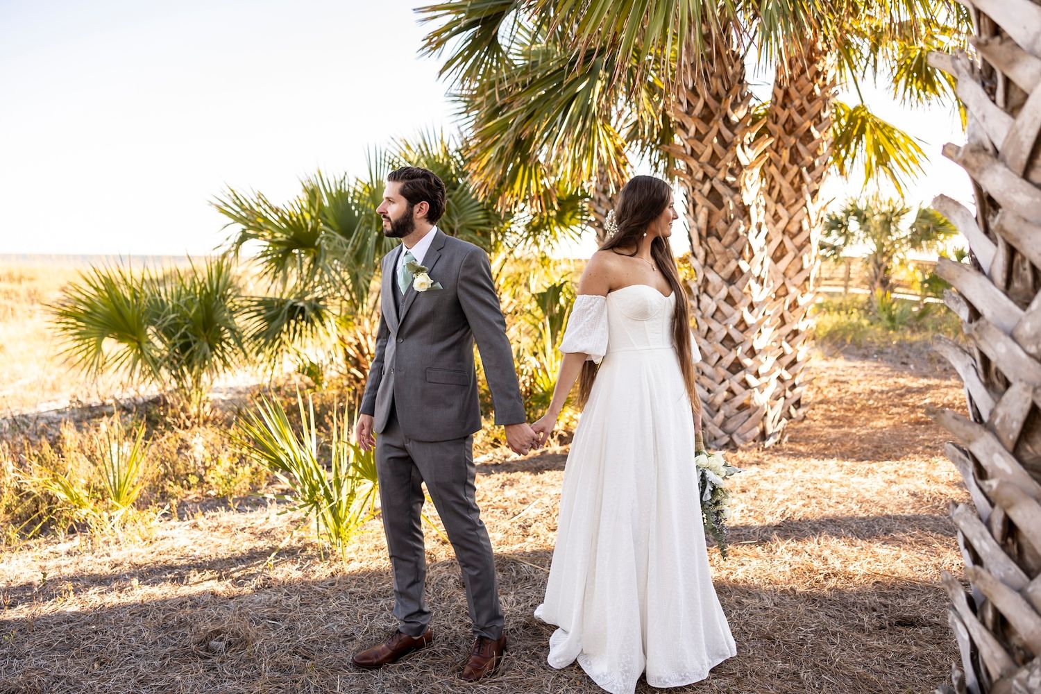 Newlywed couple stands by palm trees at sunset, bride in flowing white dress and groom in dark suit.